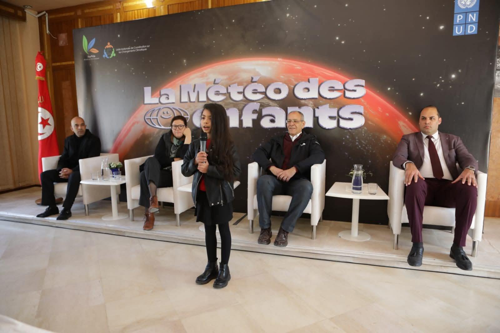 A girl speaks at a conference, with five seated individuals in the background.