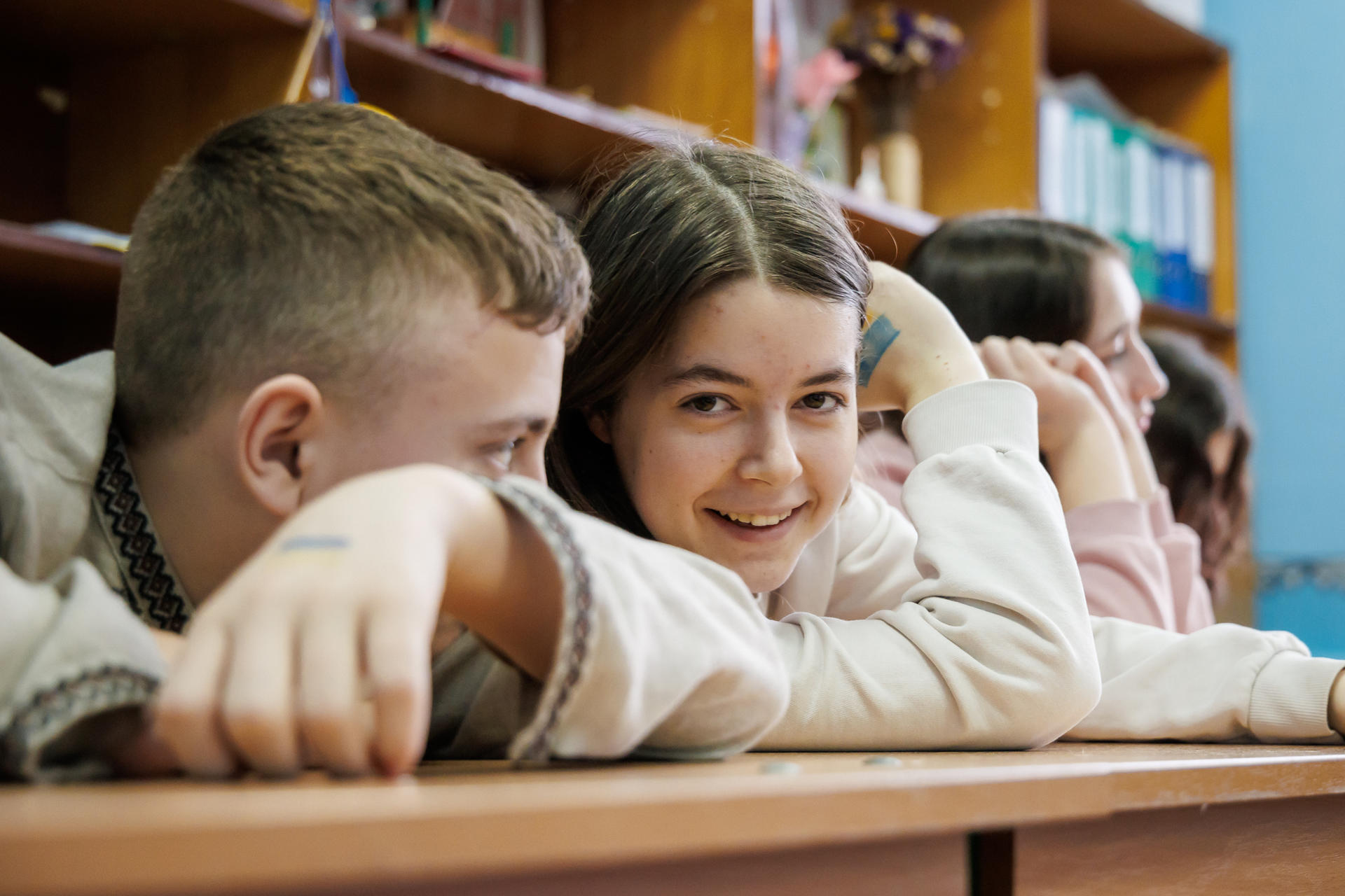 Two students smiling while leaning on a desk in a classroom, with shelves in the background.