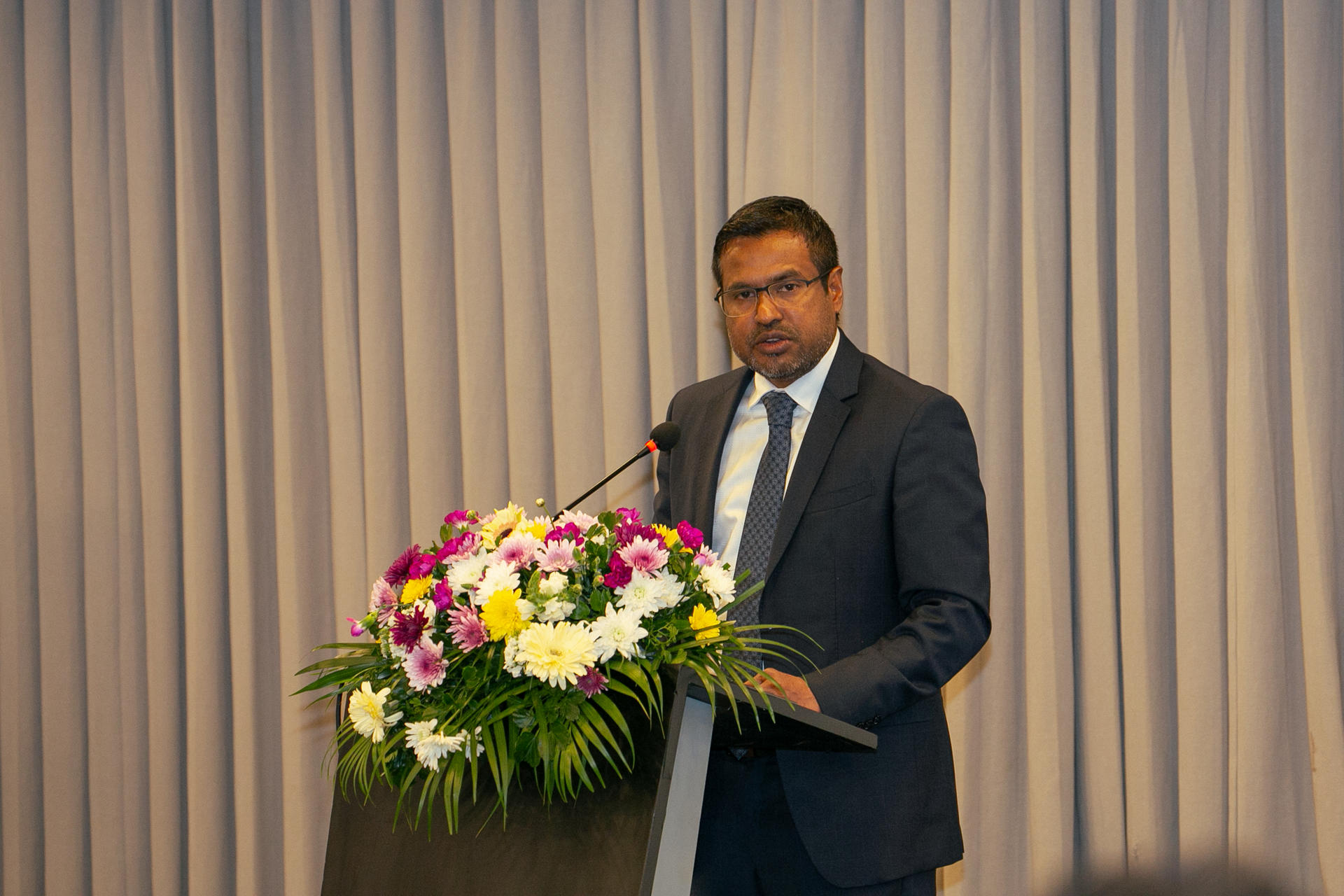 A man in a suit speaks at a podium adorned with colorful flowers.