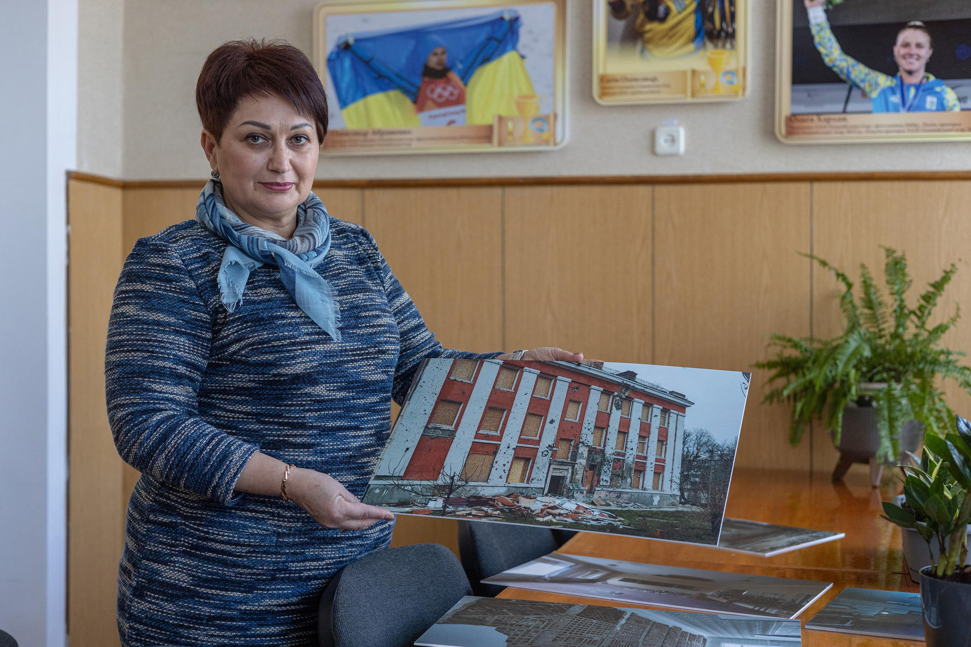 A woman in a blue sweater holds a photo of a building, surrounded by plants and framed images.