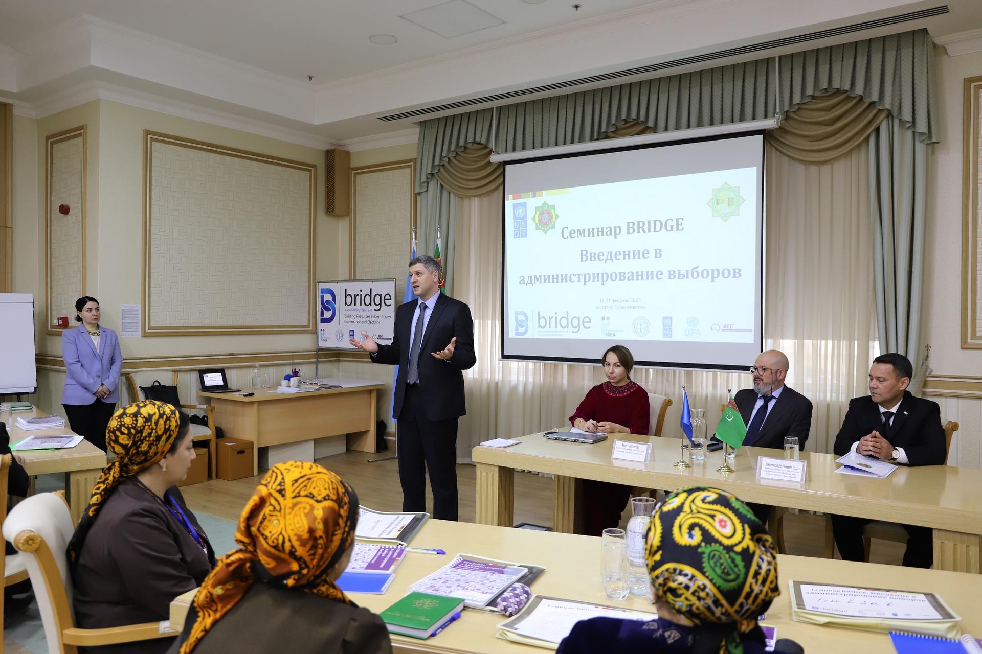 A speaker addresses an audience in a conference room, with several people attentively watching.
