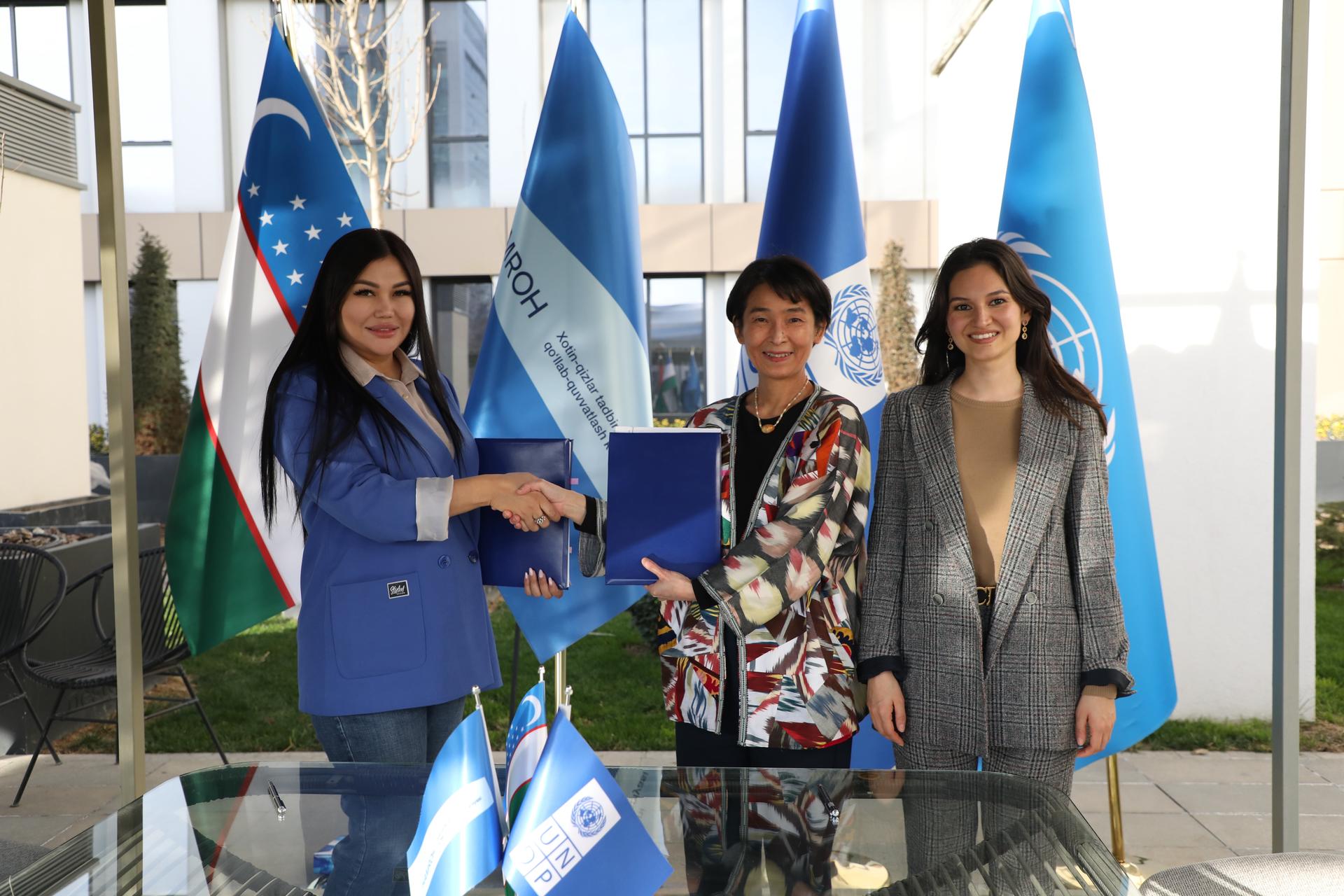 Three women smiling and shaking hands, with flags in the background.