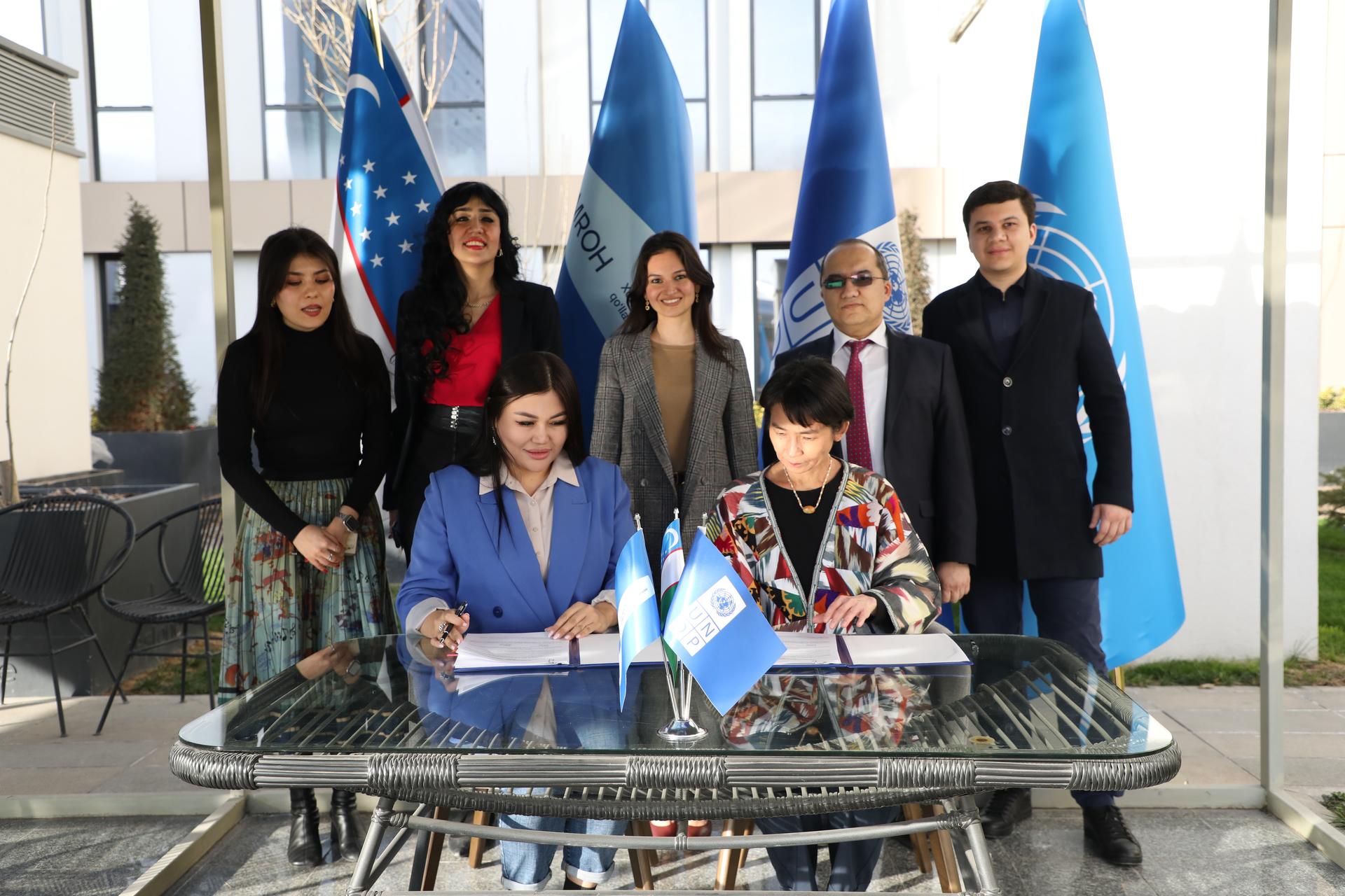 Two women sit at a table signing documents, with officials and flags in the background.