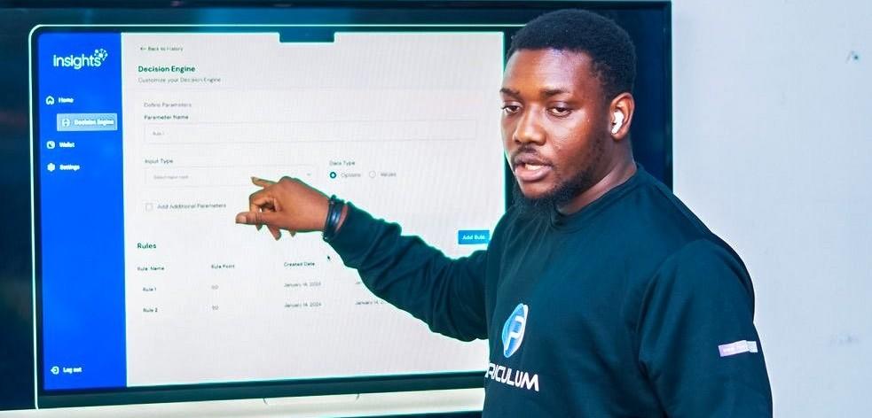A man in a black sweatshirt gestures towards a digital screen displaying data.