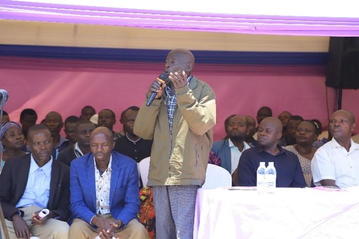 A man speaking into a microphone at a community gathering, seated audience in the background.