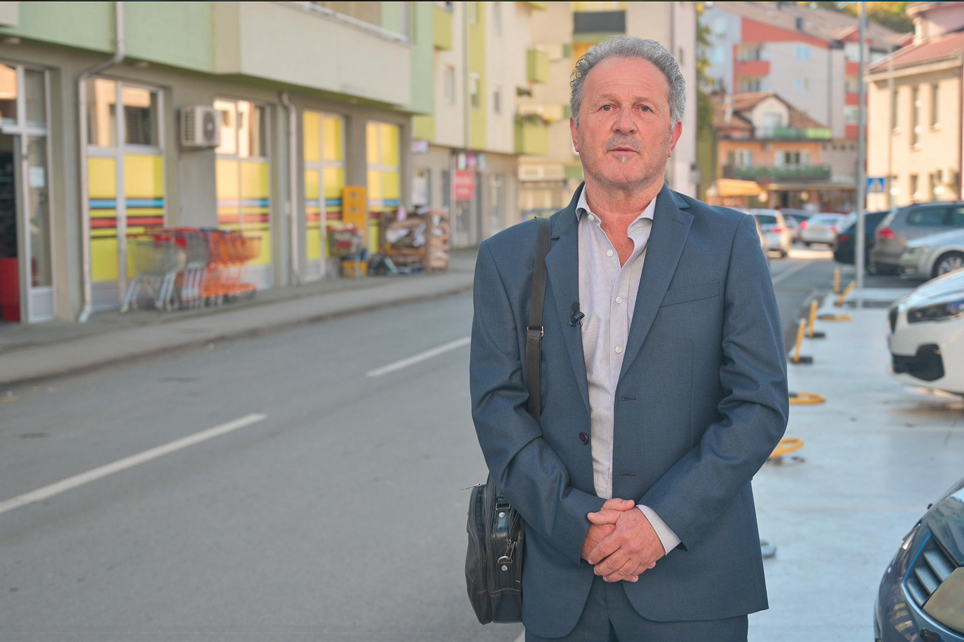 A man in a suit stands on a street with shops and parked cars in the background.