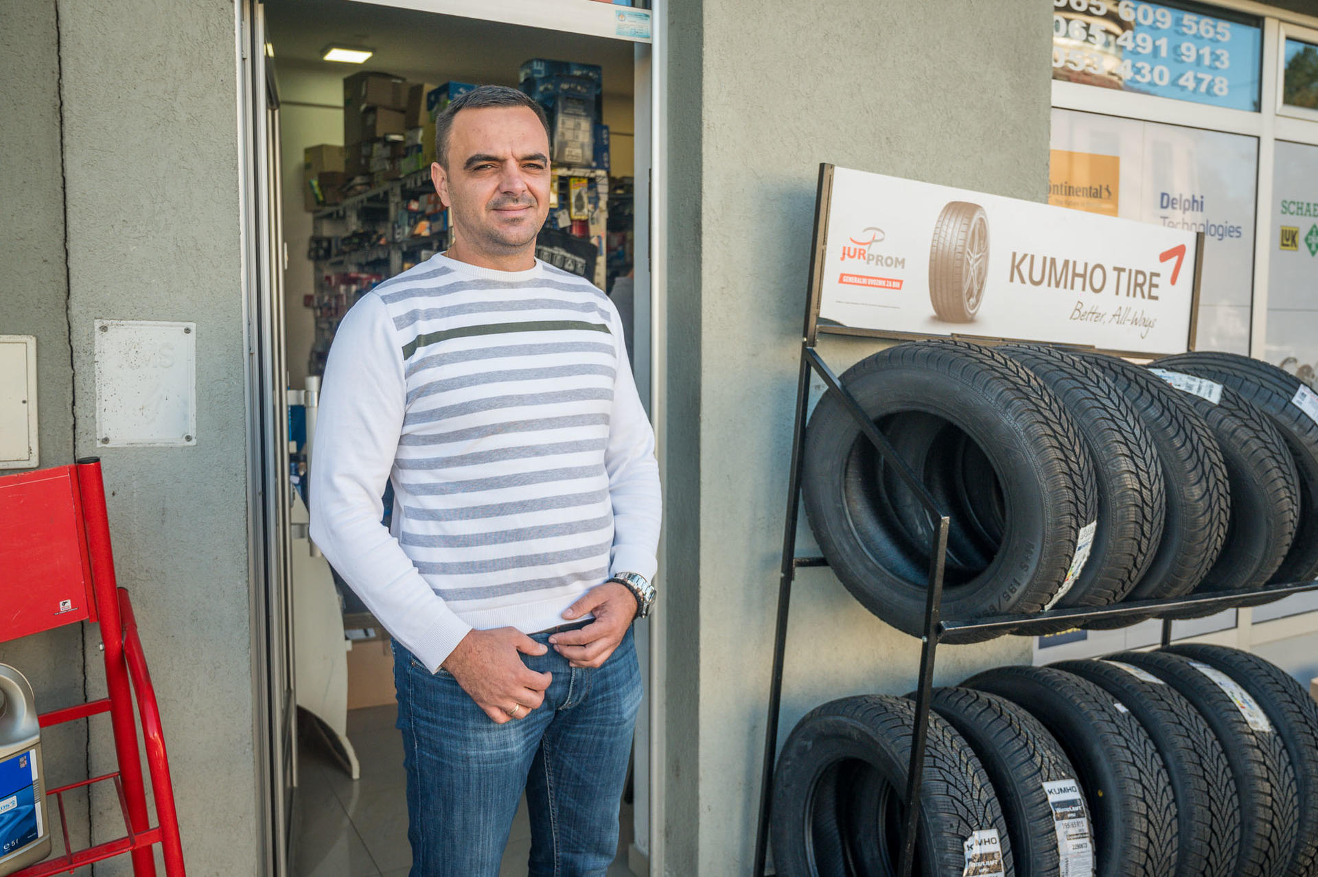 Man standing at the entrance of a tire shop, smiling, with tires displayed beside him.