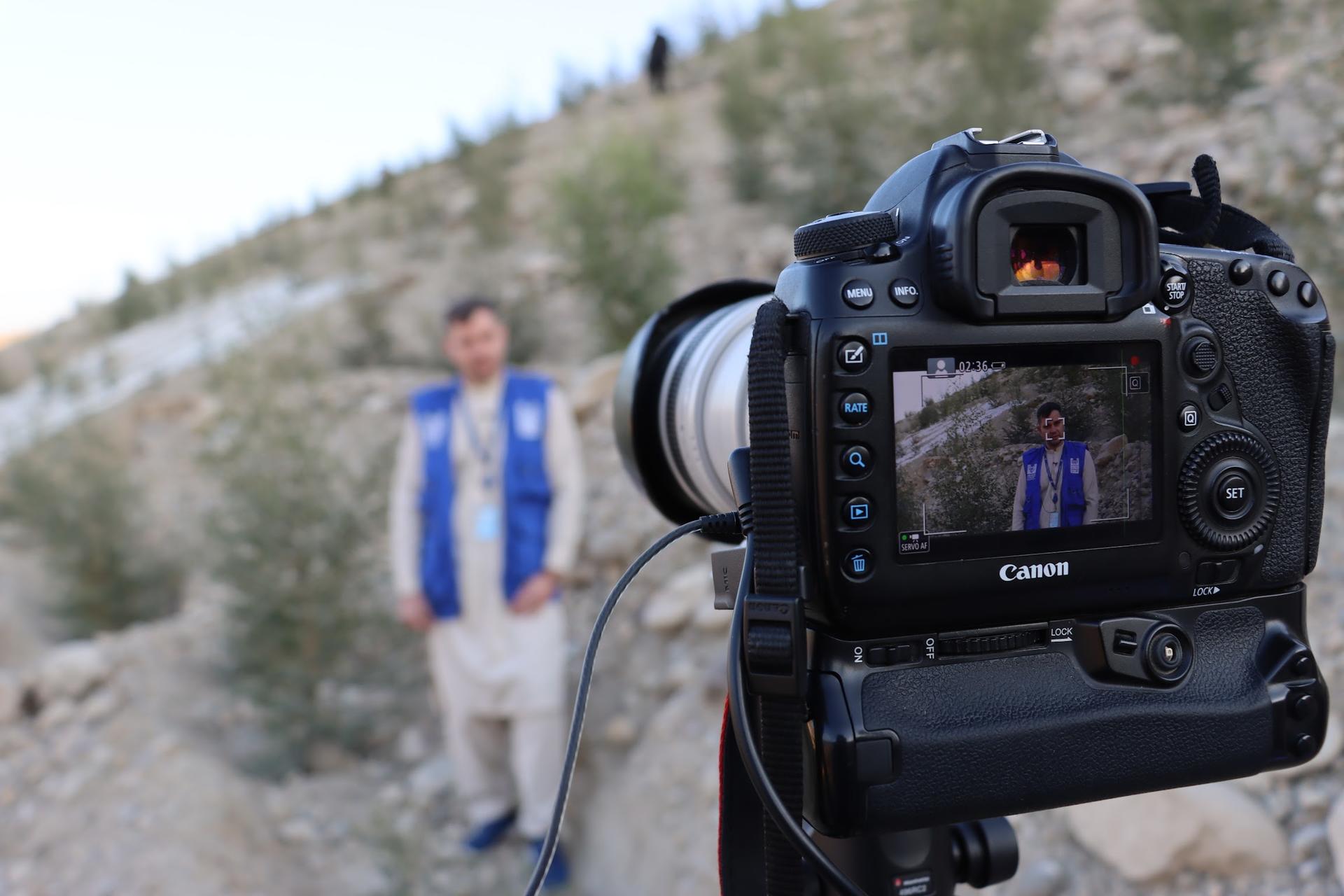 A camera captures a man in traditional clothing standing on rocky terrain.