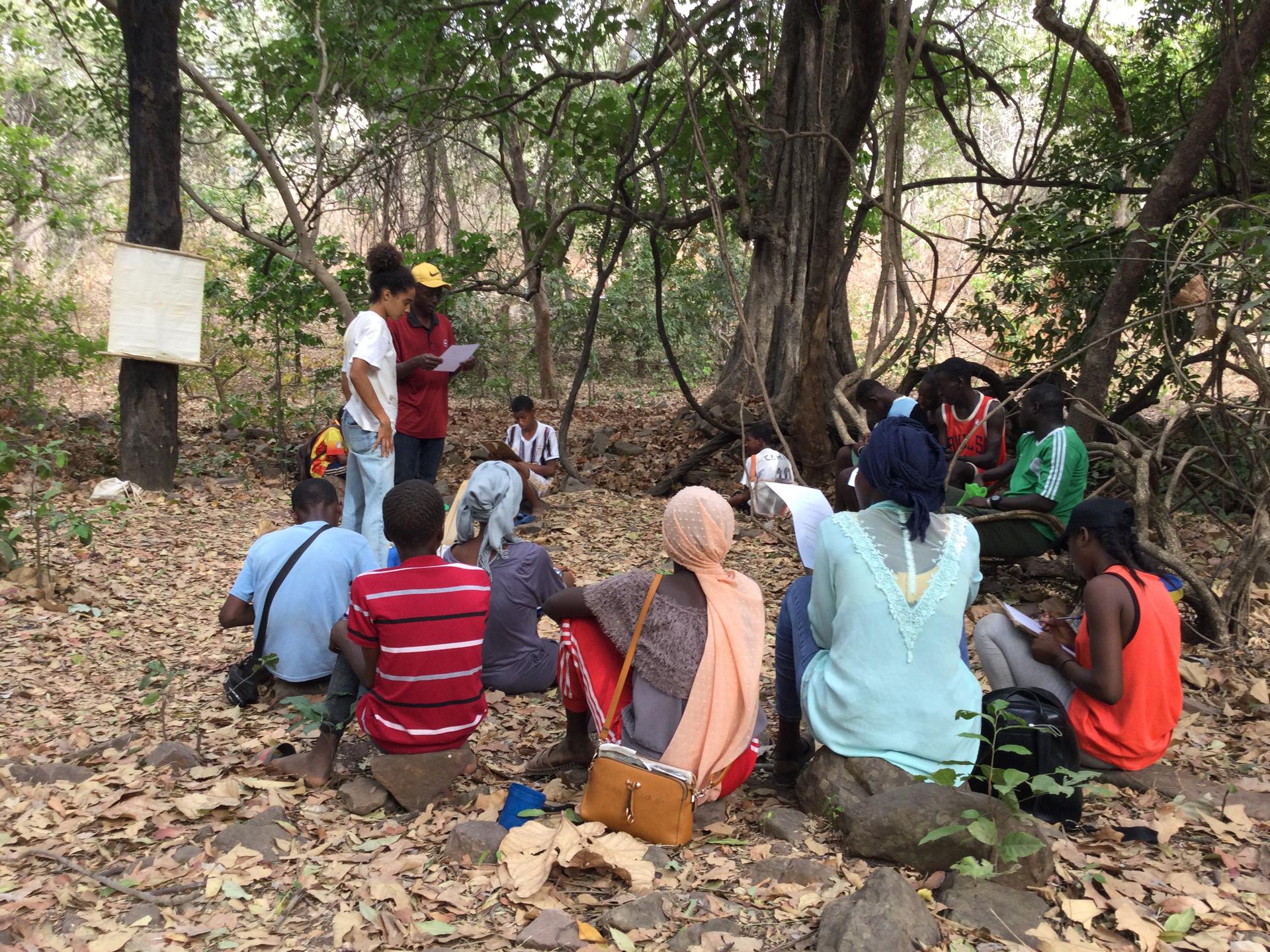 A group of people sitting on the ground in a wooded area, listening to a speaker.