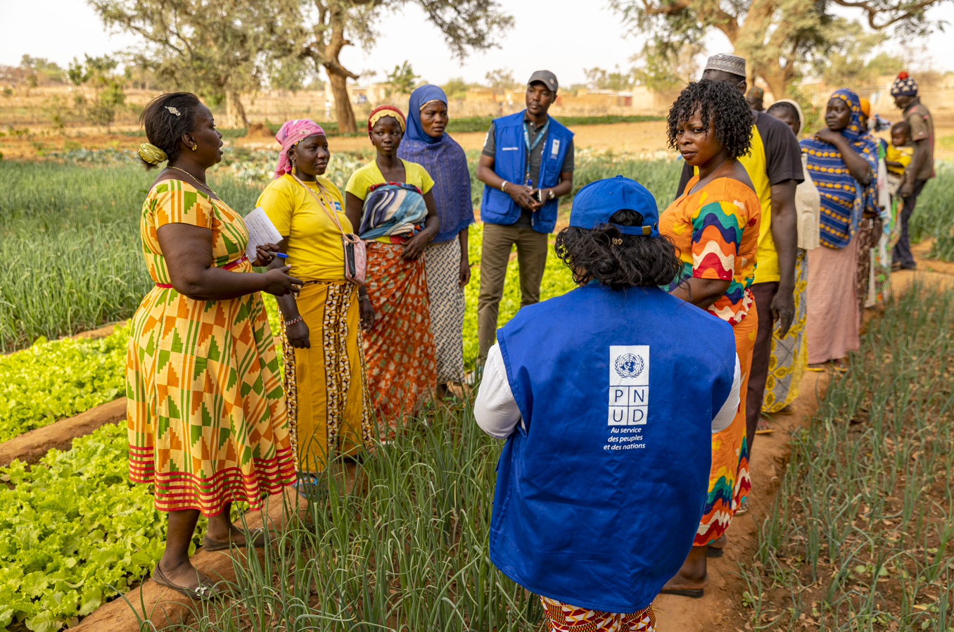 A group of people in colorful clothing engaged in a discussion in a green farm area.