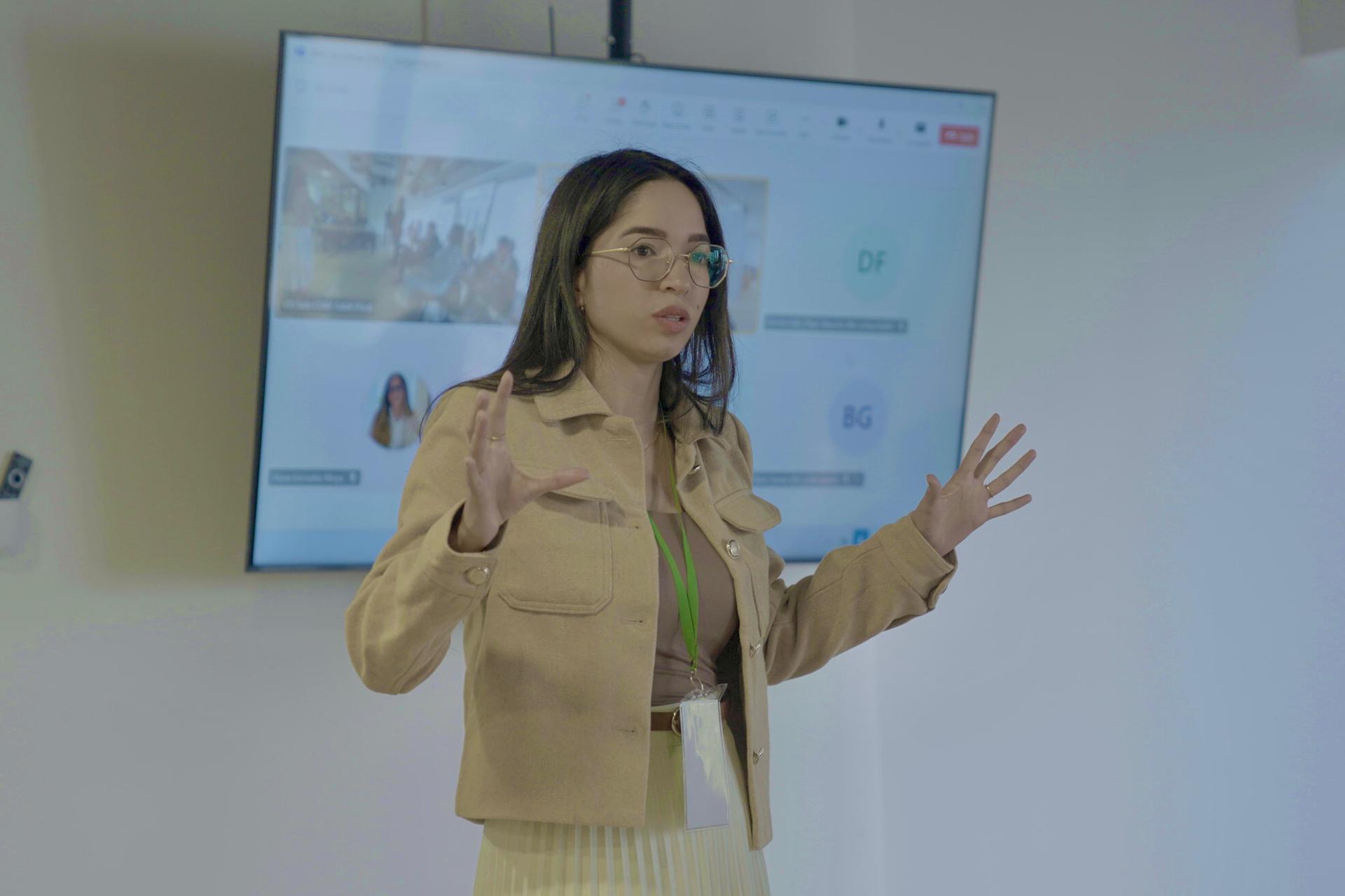 A woman gestures while speaking in front of a presentation screen in a modern setting.