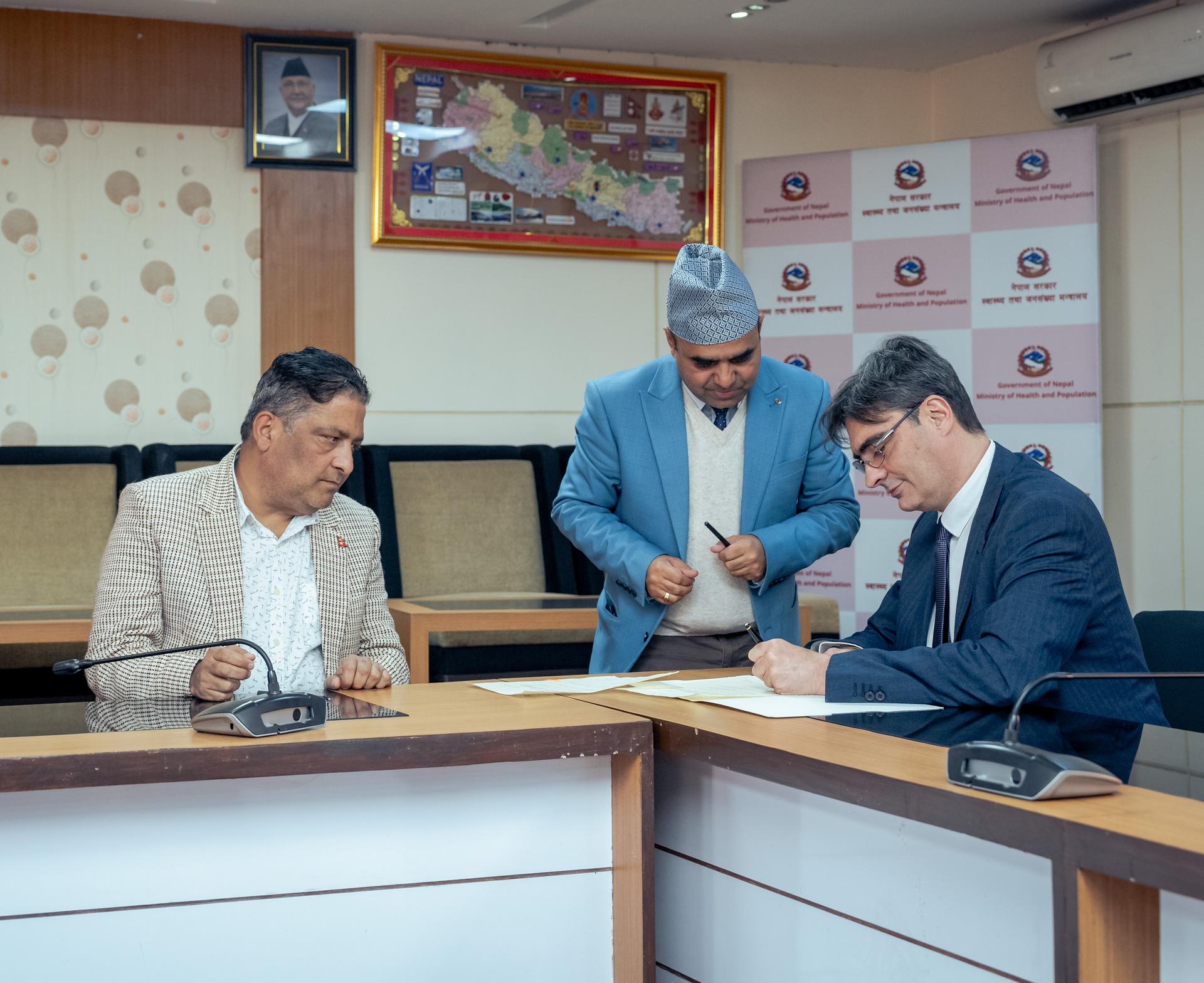 Two men sit at a table signing documents, with a third man standing and observing.