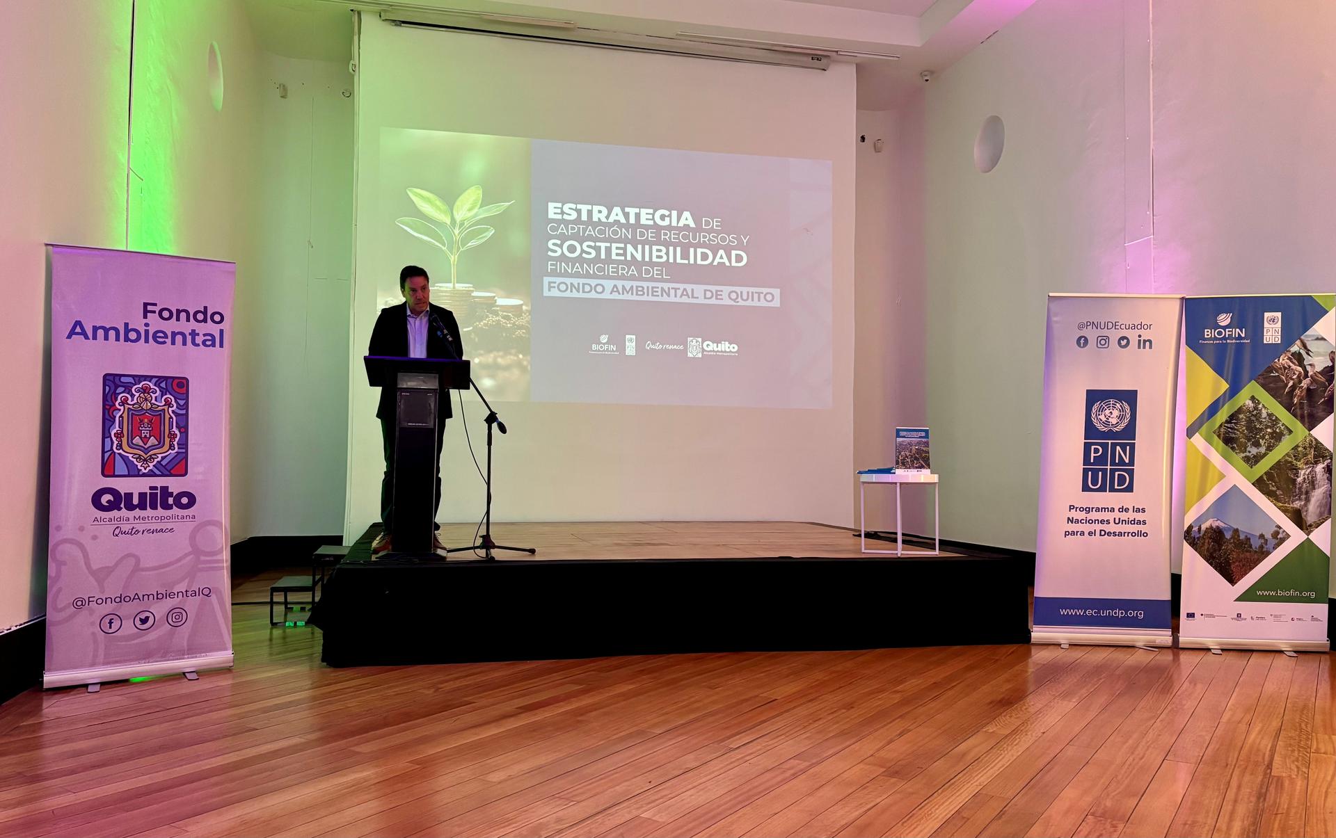 A speaker presents on a stage, with displayed banners and green lighting in a spacious room.