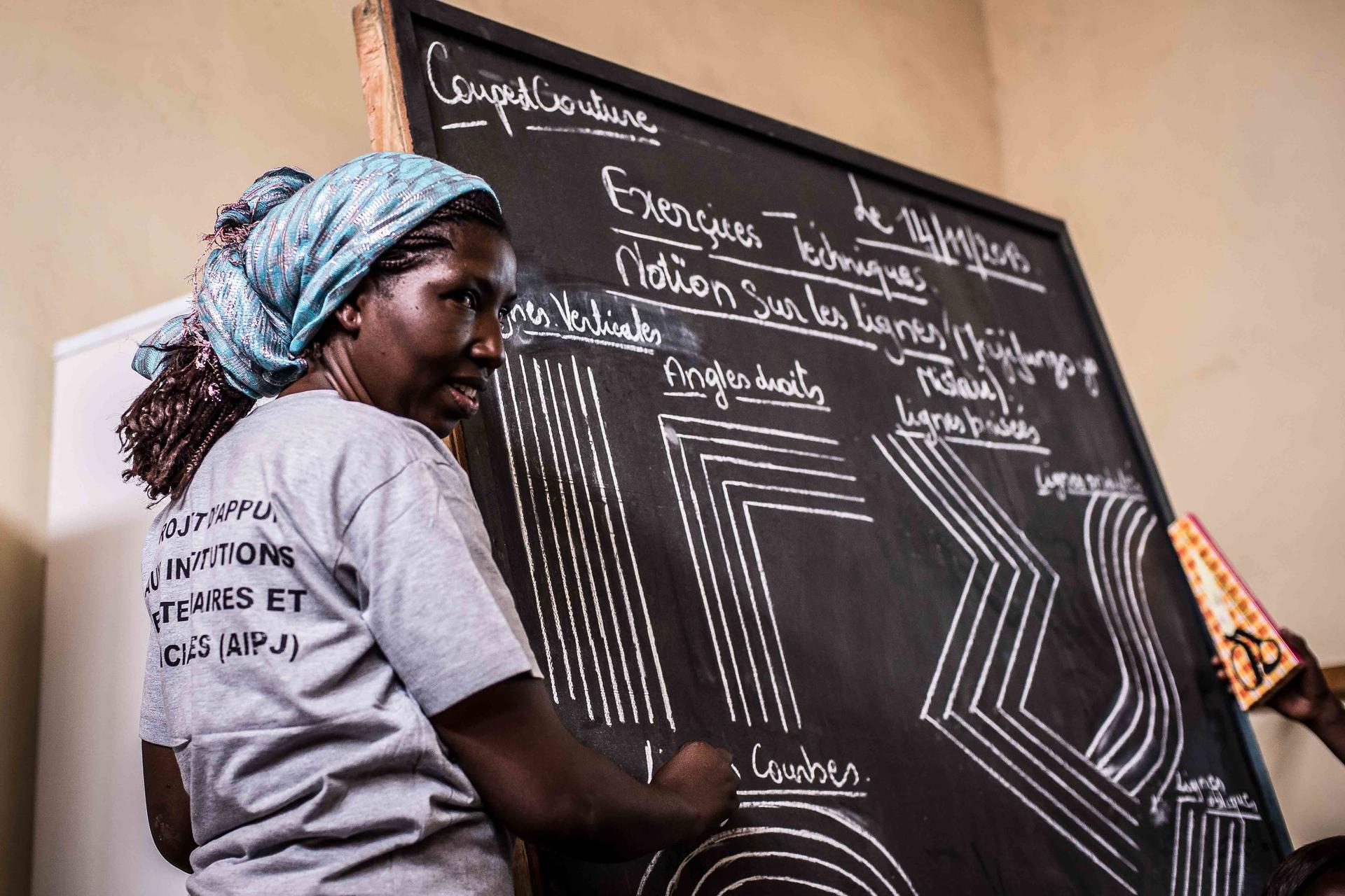 Woman with a headscarf teaches at a chalkboard filled with patterns and text.