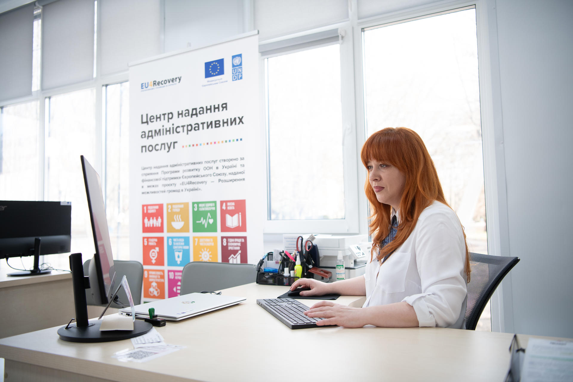 A woman with red hair works at a desk in an office setting, focused on her computer.