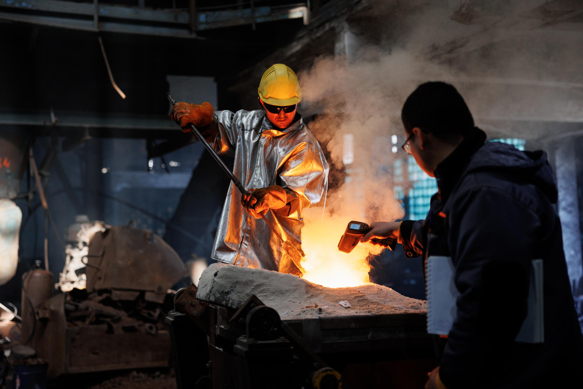 Workers in a foundry, one pouring molten metal, the other observing with a flashlight.