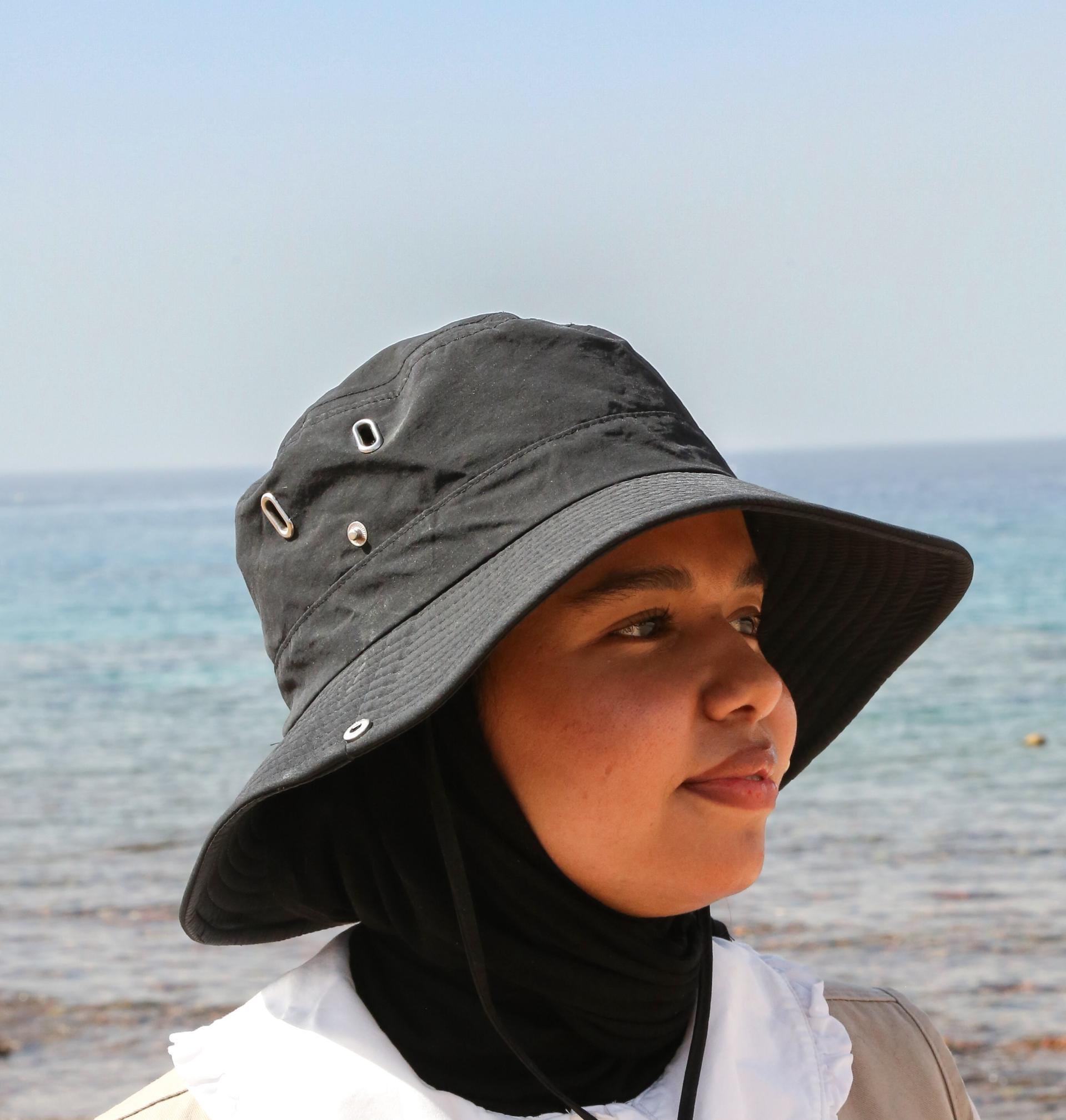 A woman wearing a black bucket hat and black hijab, facing the sea.