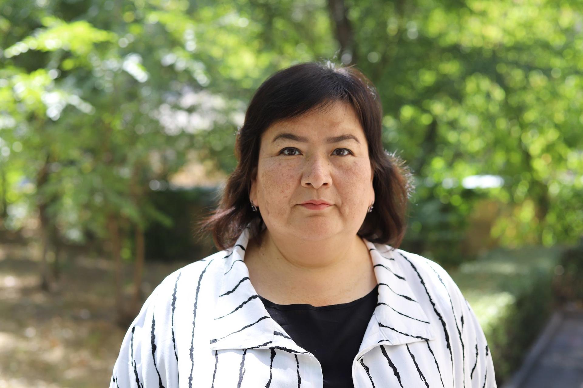 A woman with dark hair stands outdoors, wearing a striped white and black jacket, surrounded by greenery.
