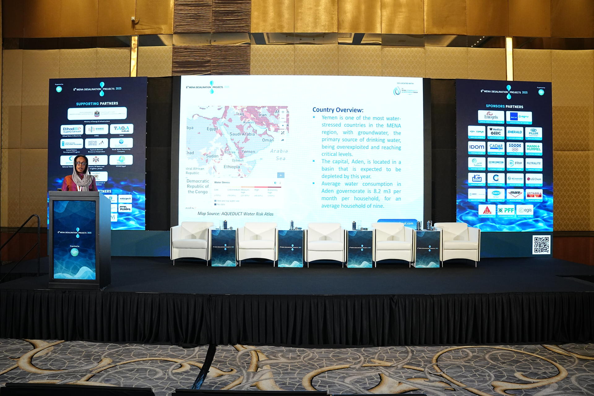 A speaker presents at a conference with a large screen displaying a map and data.