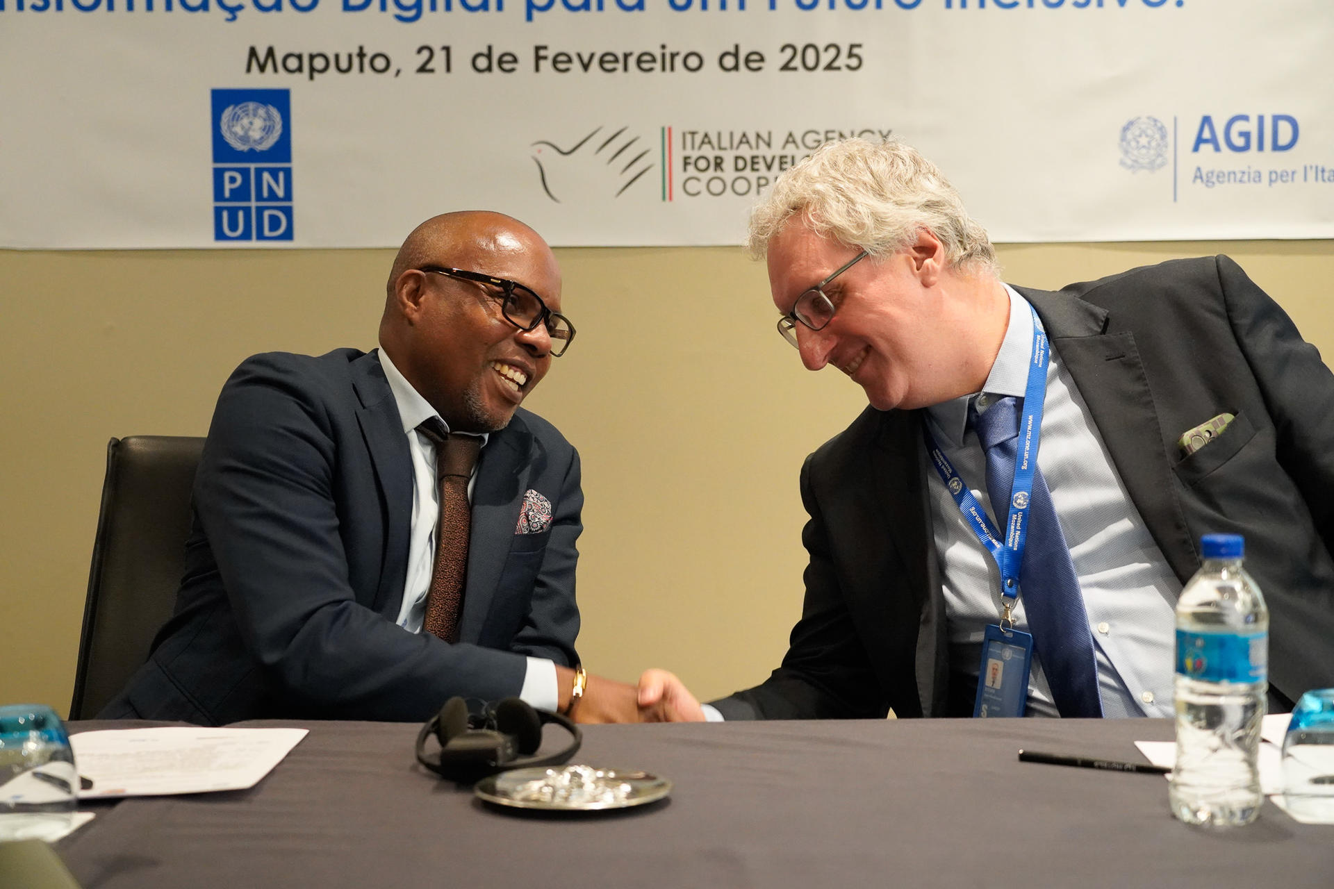 Two men shaking hands at a conference table, smiling, with water bottles and papers visible.