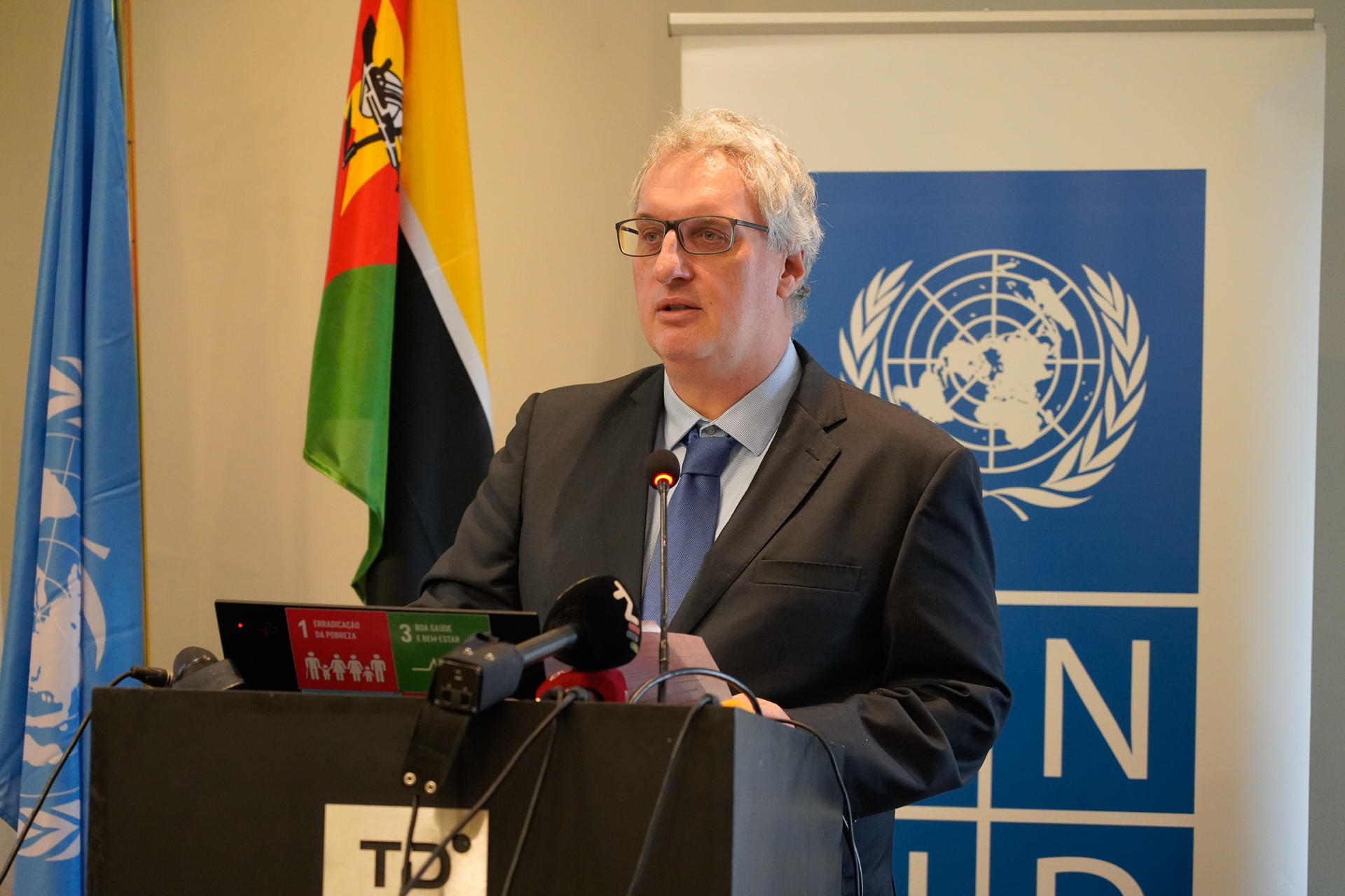 A man in a suit speaks at a podium with UN and national flags in the background.