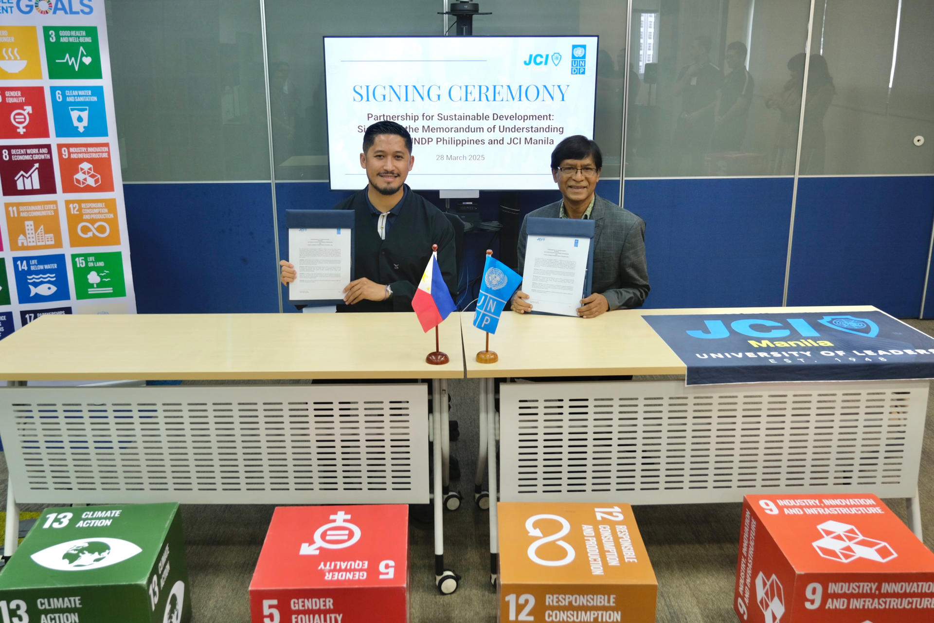 Two men hold signed documents at a table, with colorful boxes and a presentation screen behind.