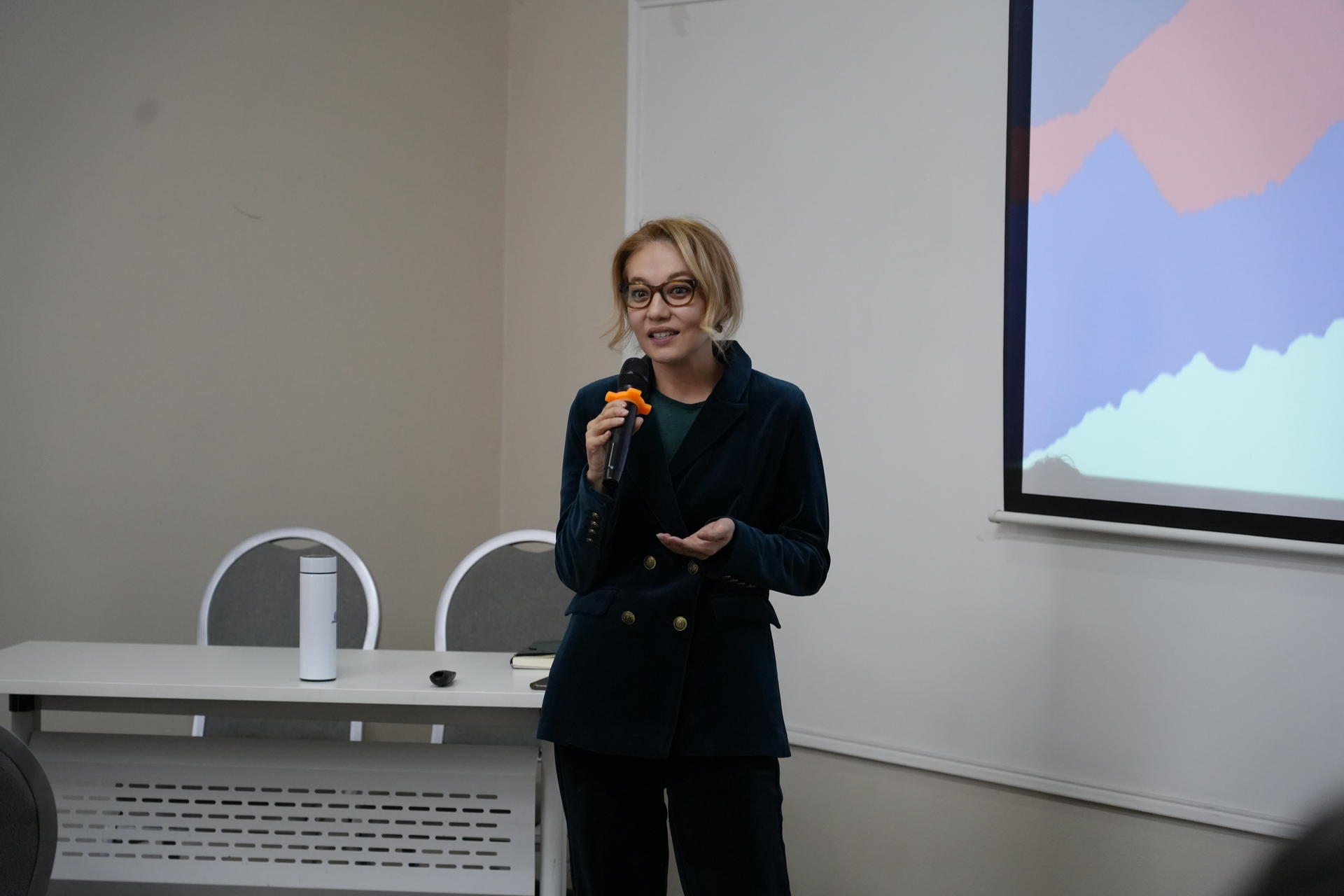 A woman in a dark blazer speaks into a microphone in front of a projector screen.