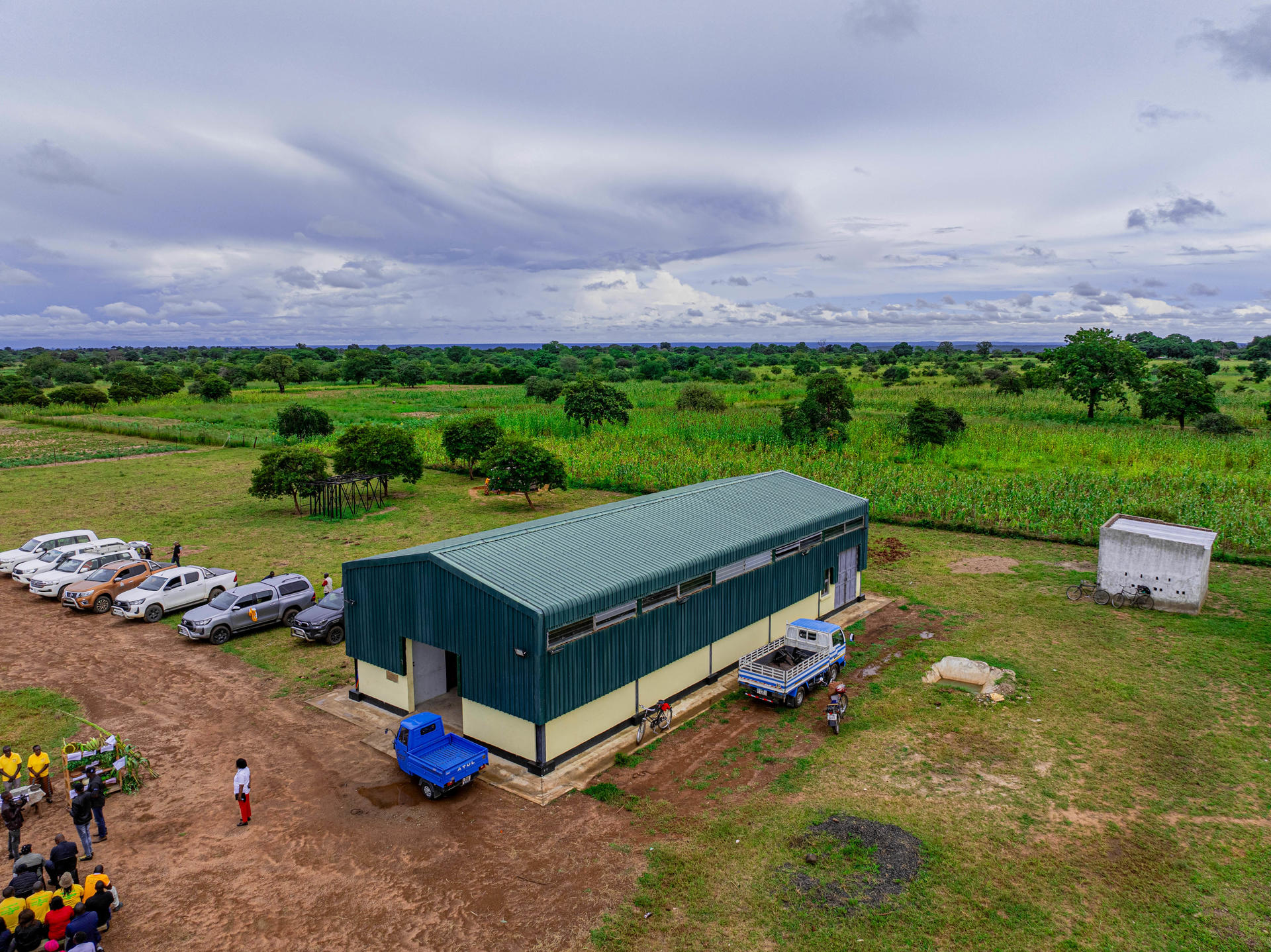 A green agricultural building with cars parked nearby, set against a grassy landscape and cloudy sky.