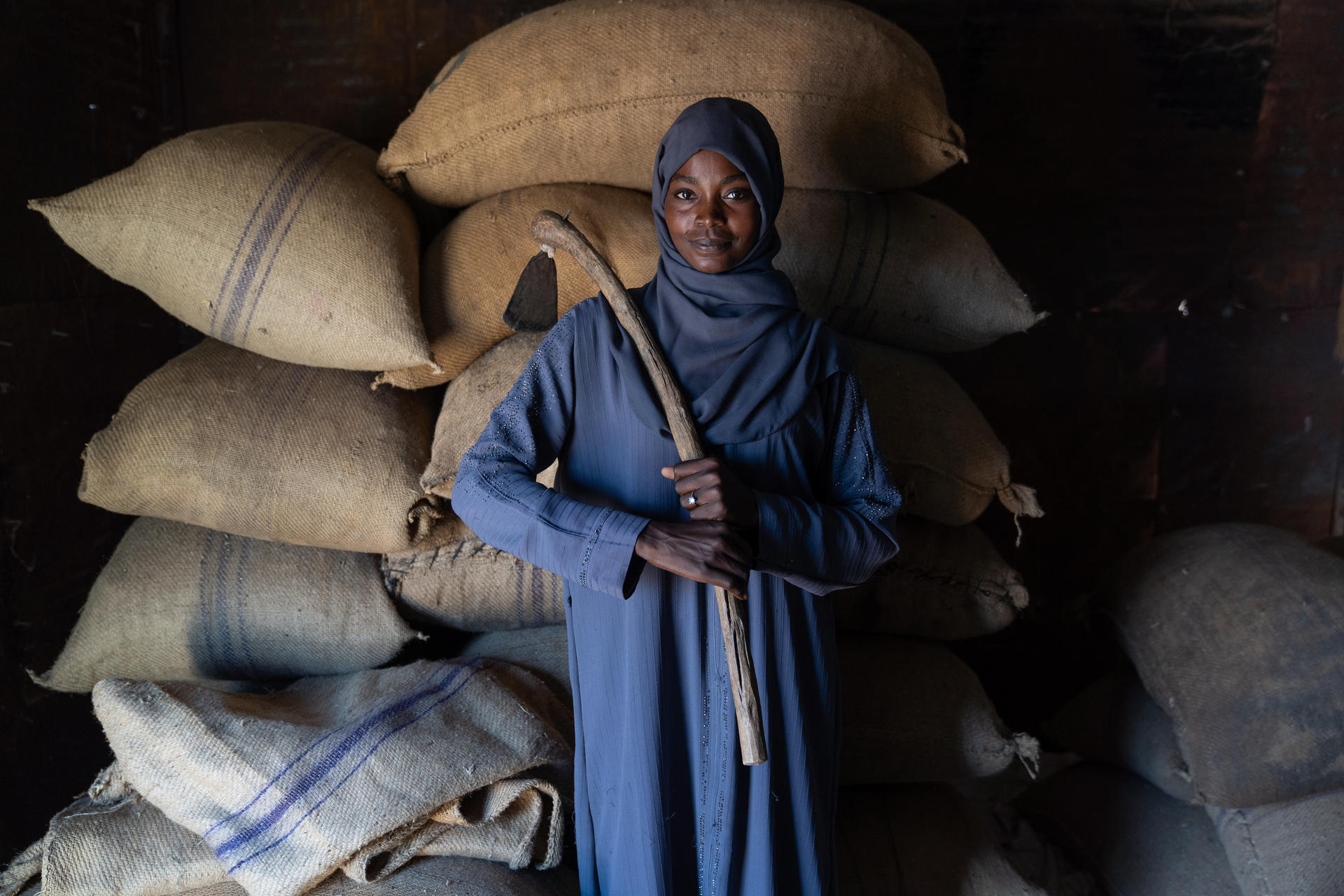 A woman in a gray dress stands smiling with a stick, surrounded by large sacks.