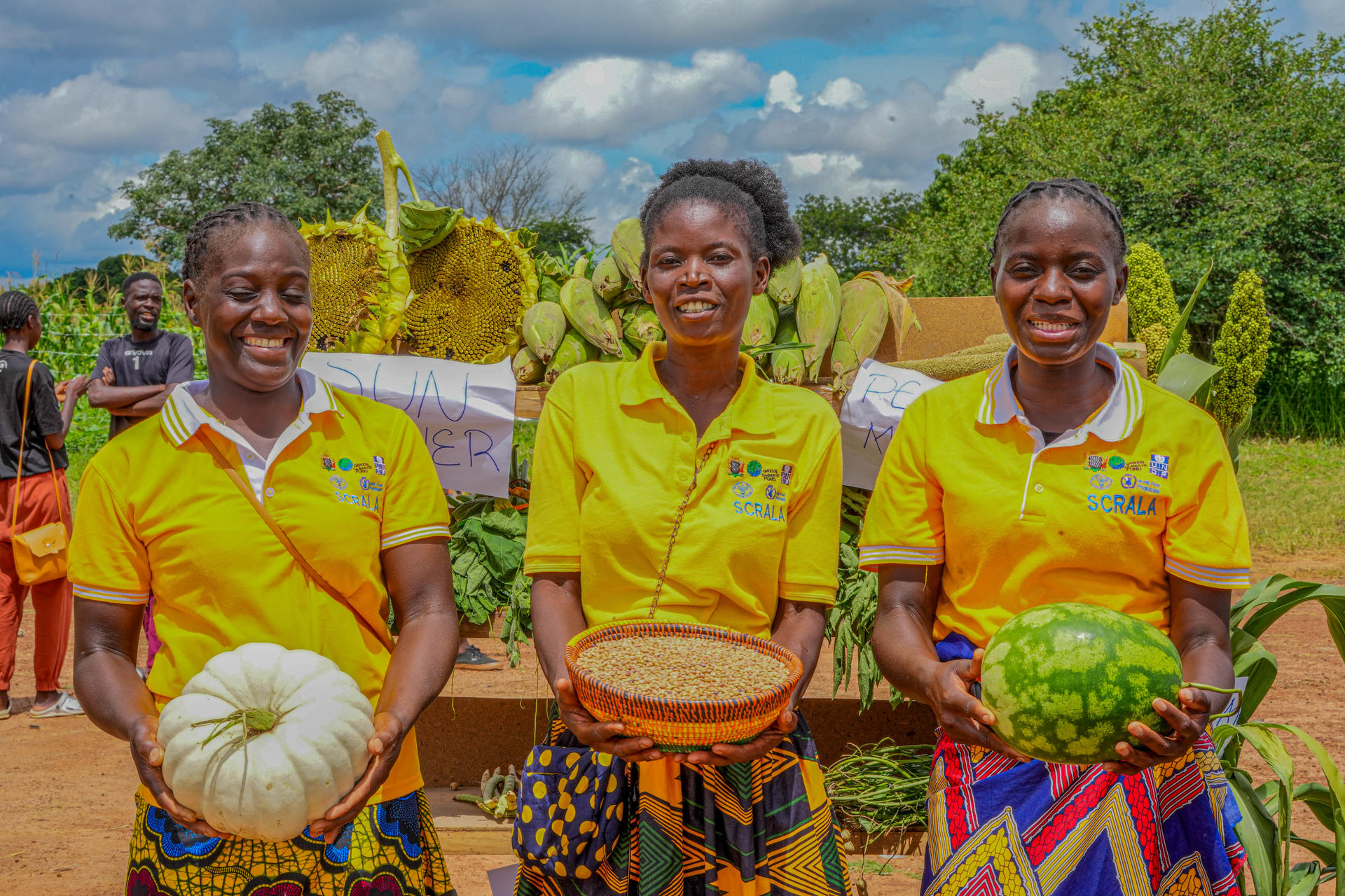 Three women in yellow shirts smile while holding a pumpkin, basket of grains, and watermelon.