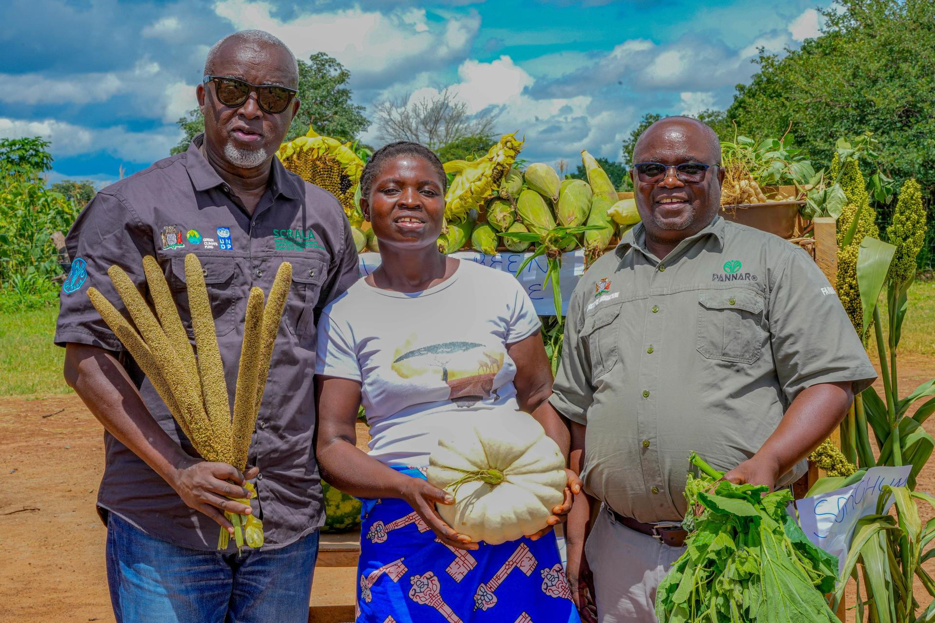Three people stand in a field, holding vegetables, with a backdrop of green crops and blue sky.