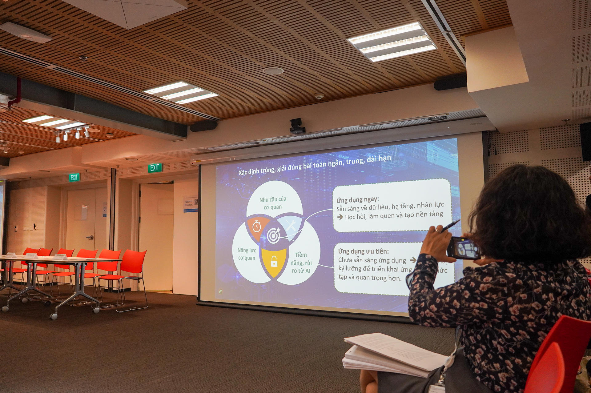 A person sits in front of a presentation screen with a Venn diagram and red chairs in the background.