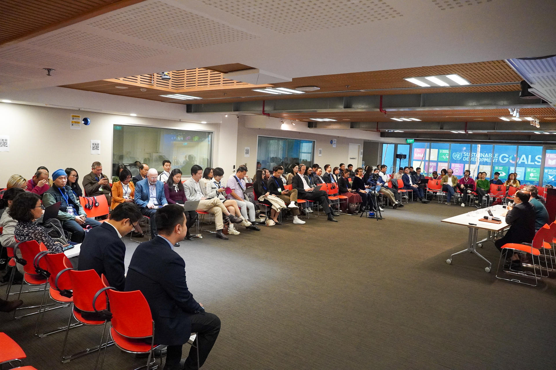 A large crowd seated in a modern room with red chairs, facing a speaker at a table.