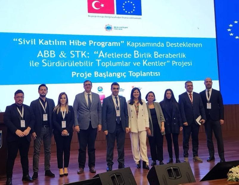 A group of professionals poses together on stage at a conference, with flags and logos in the background.