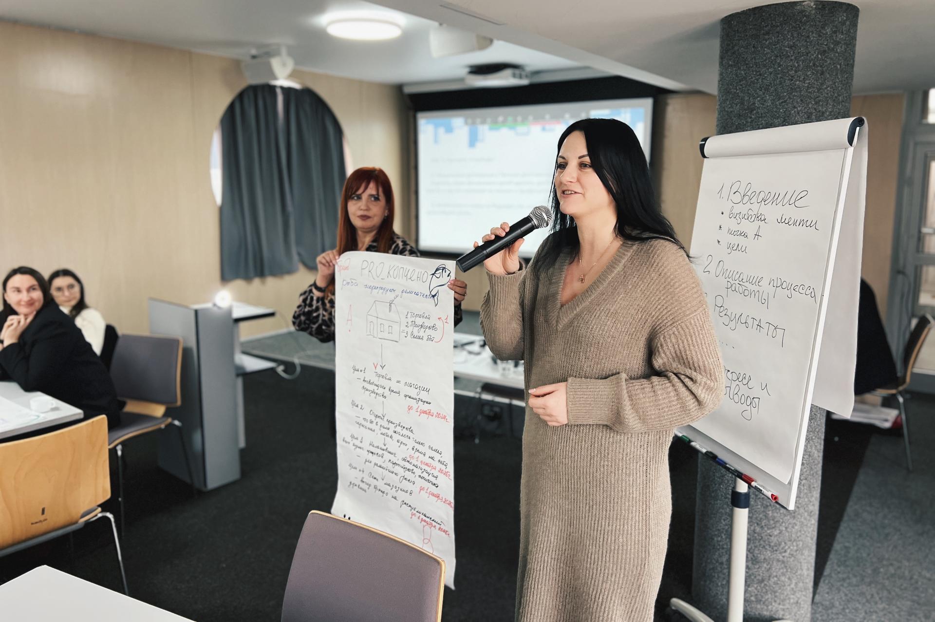 Two women presenting in a meeting room, one holding a microphone and the other a poster.