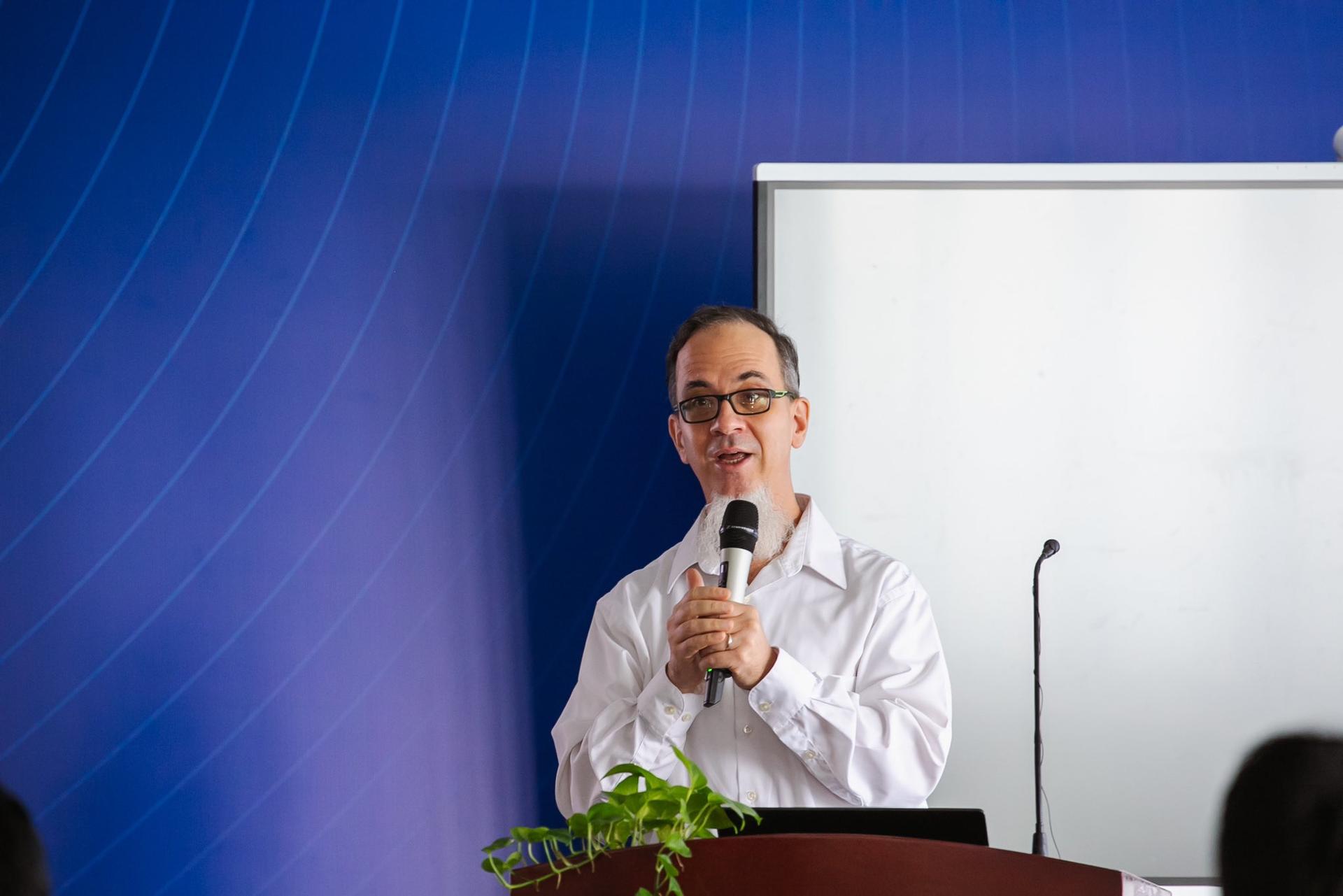 A man stands at a podium, speaking into a microphone, with a blue background.