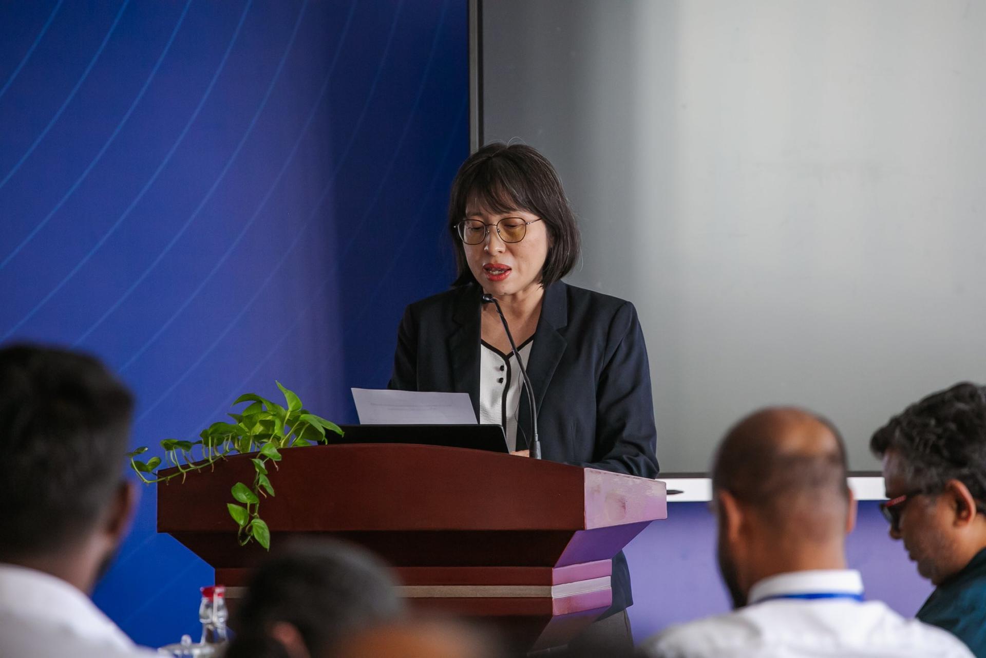 A woman speaks at a podium, addressing an audience in a conference setting.