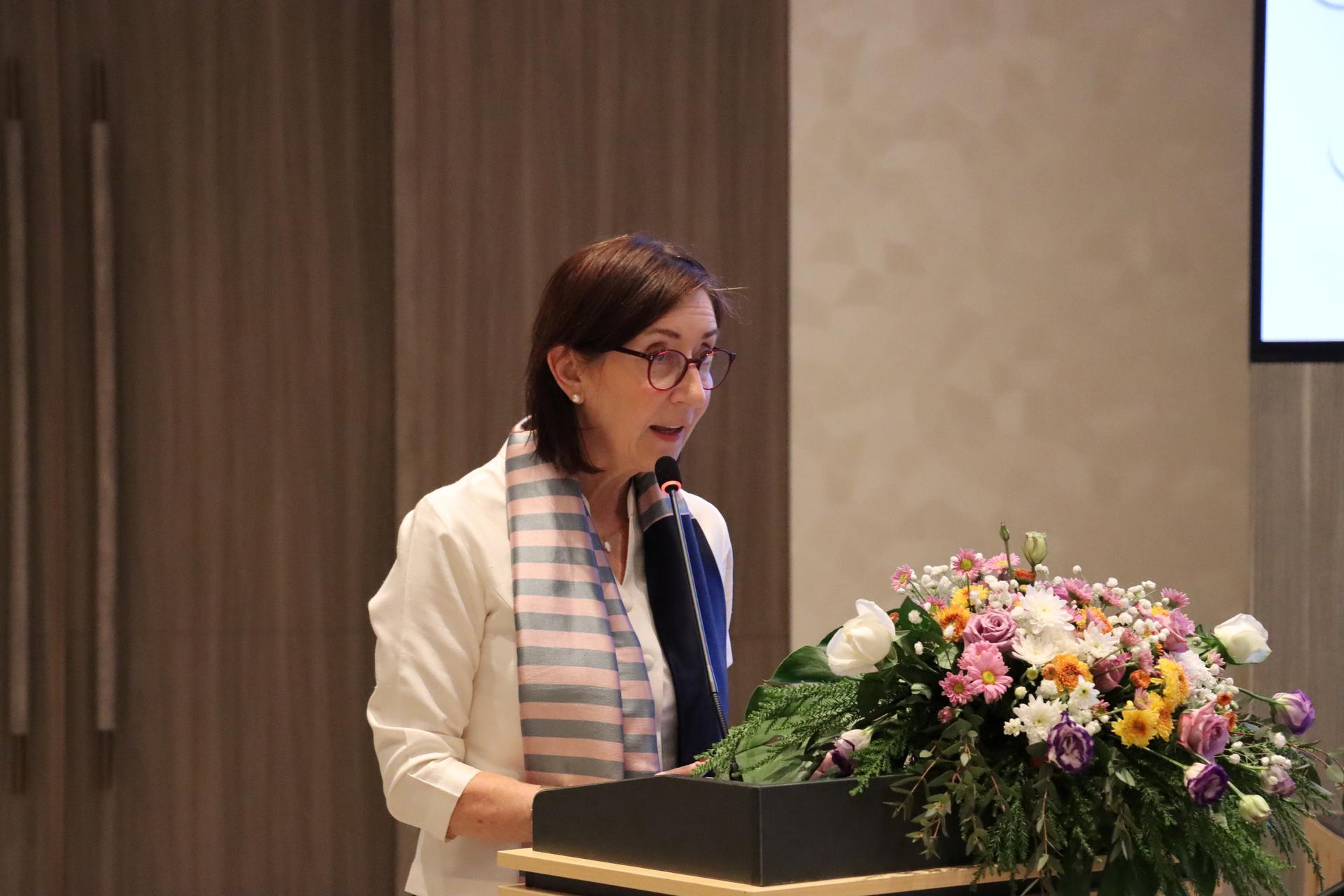 A woman speaking at a podium with a floral arrangement, dressed in a white jacket and scarf.
