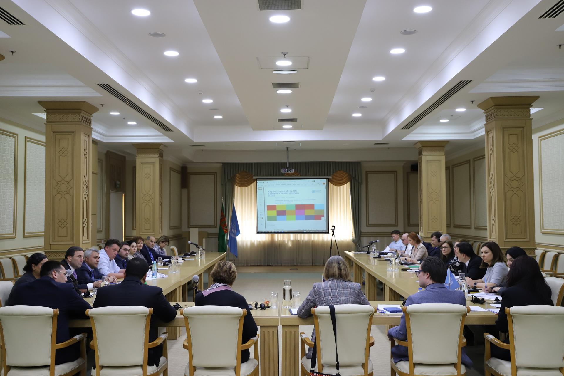 A formal meeting in a spacious room with a large table, displaying a presentation screen.