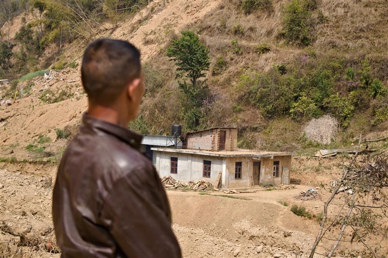 A man in a brown jacket gazes at a small, abandoned house on a hillside.