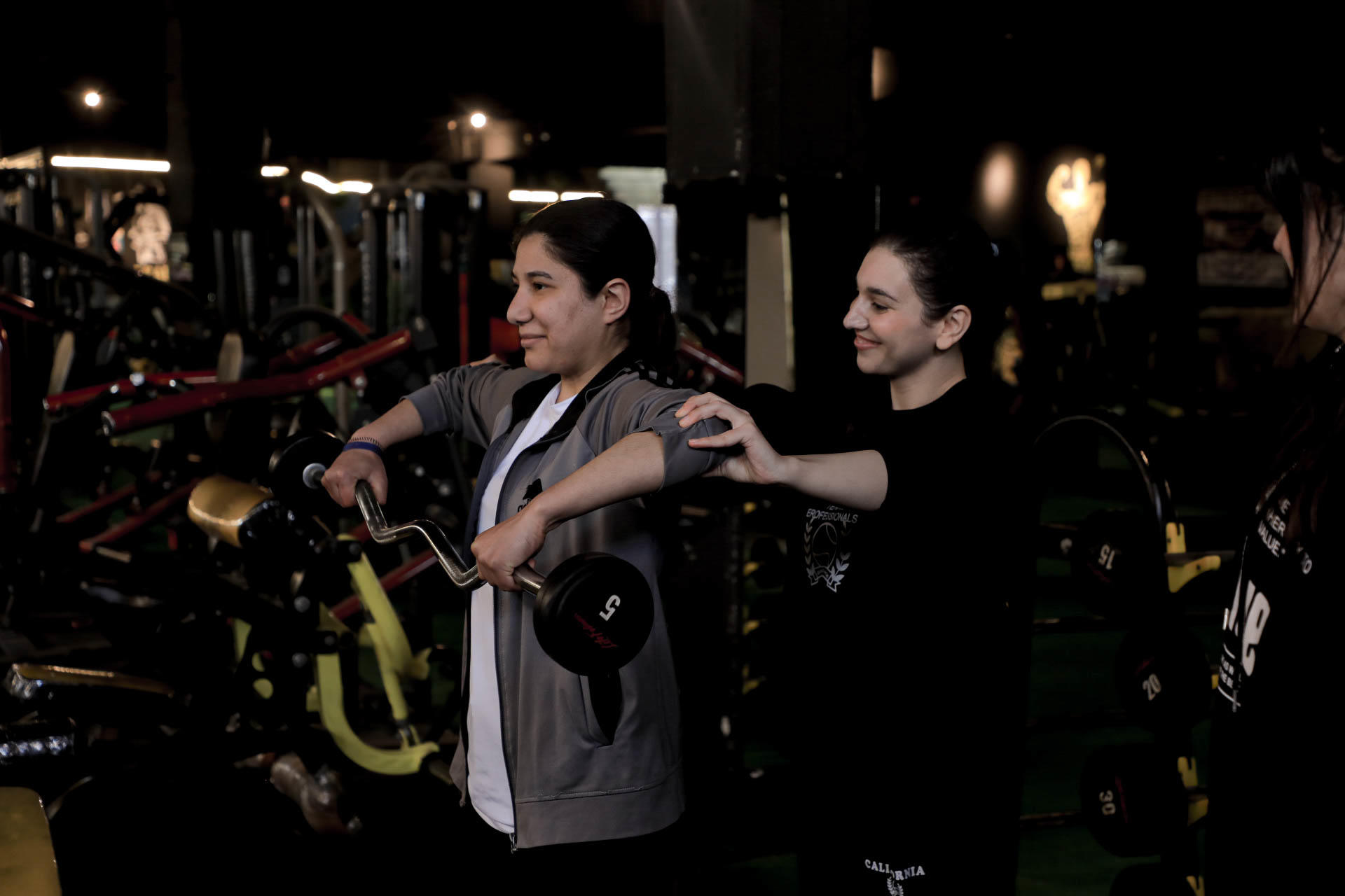 Two women exercising in a gym, one assisting the other with dumbbell weights.
