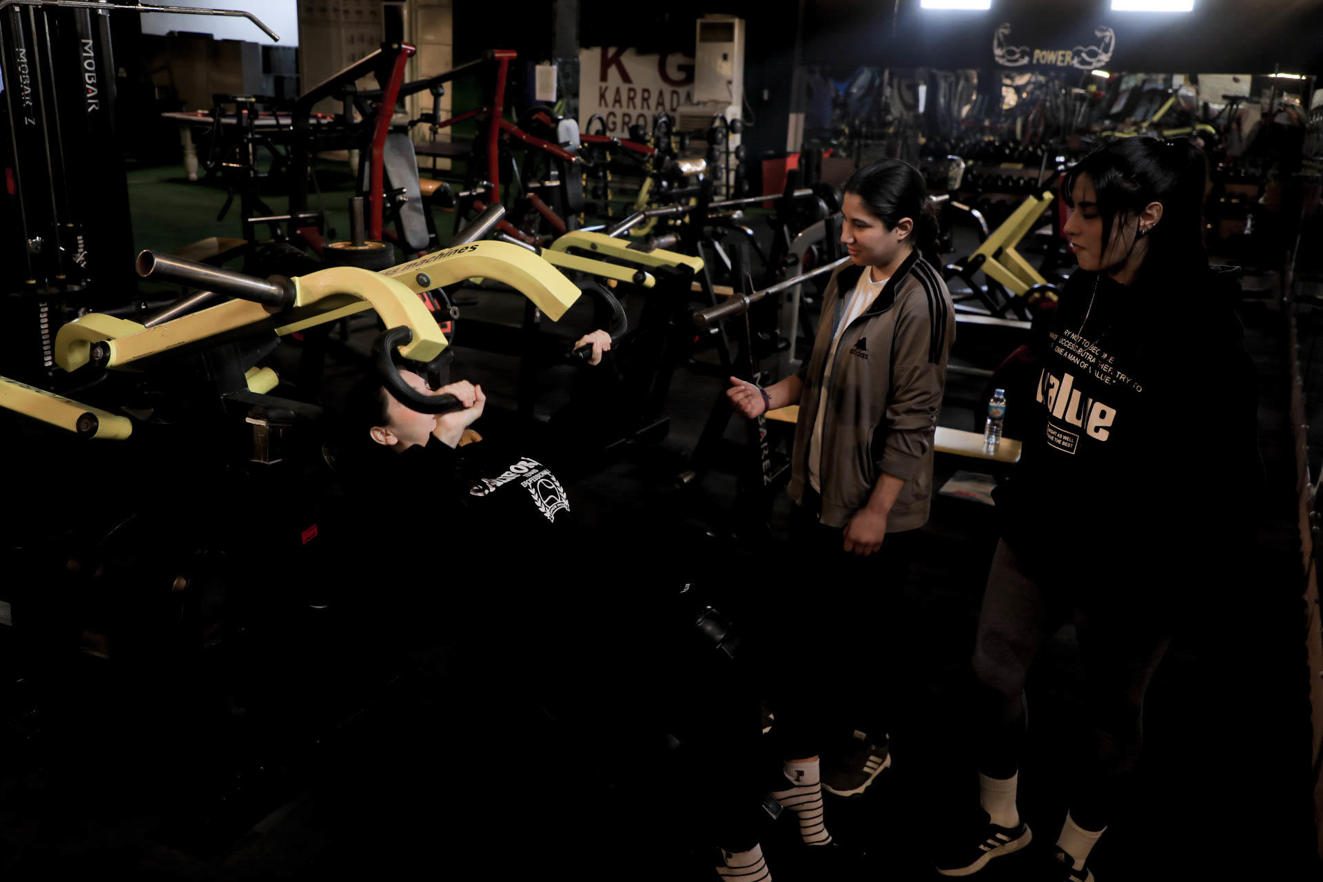 Two women assist another woman lifting weights in a dimly lit gym.
