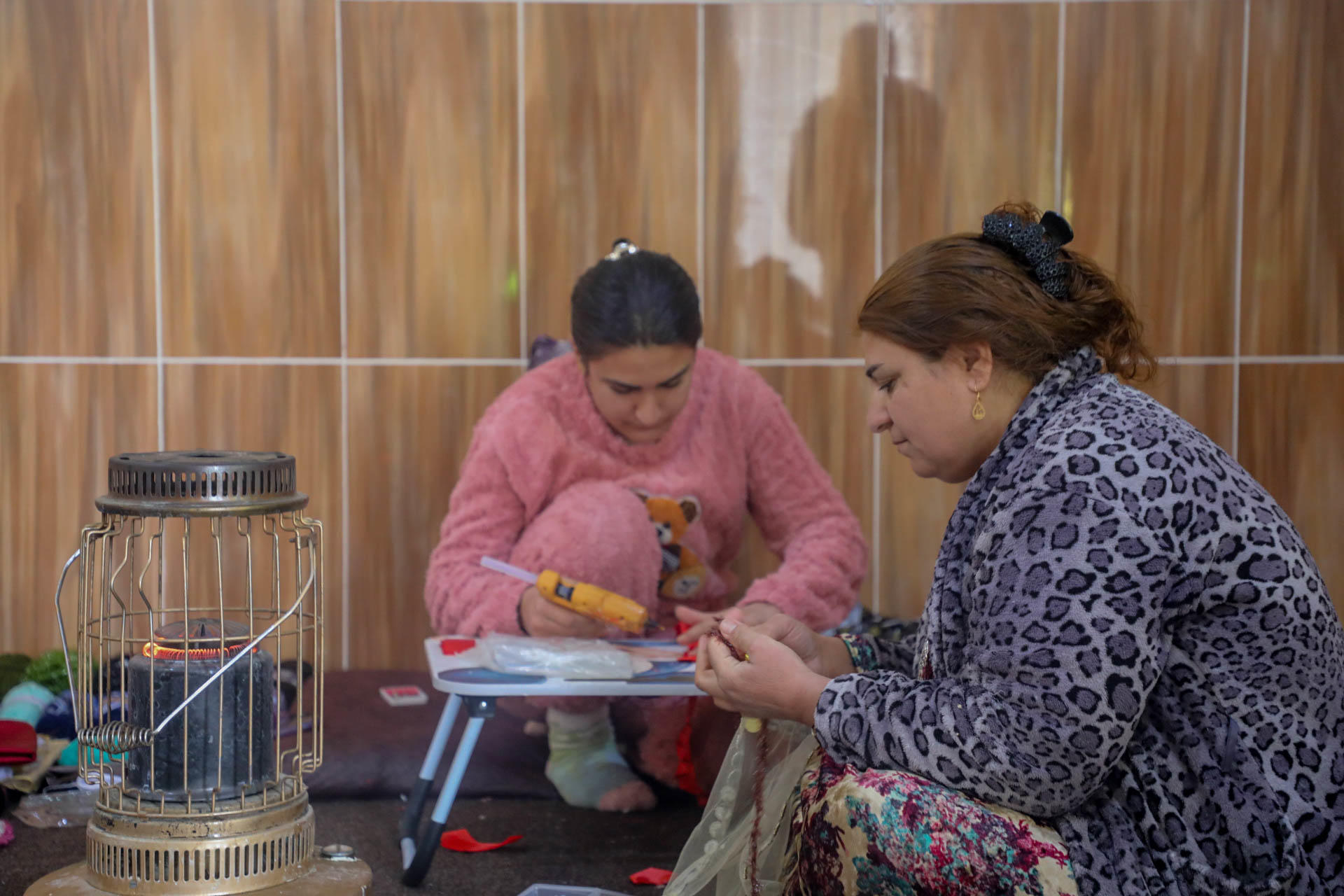 Two women working together on a table, with a stove heater nearby, in a warm interior setting.