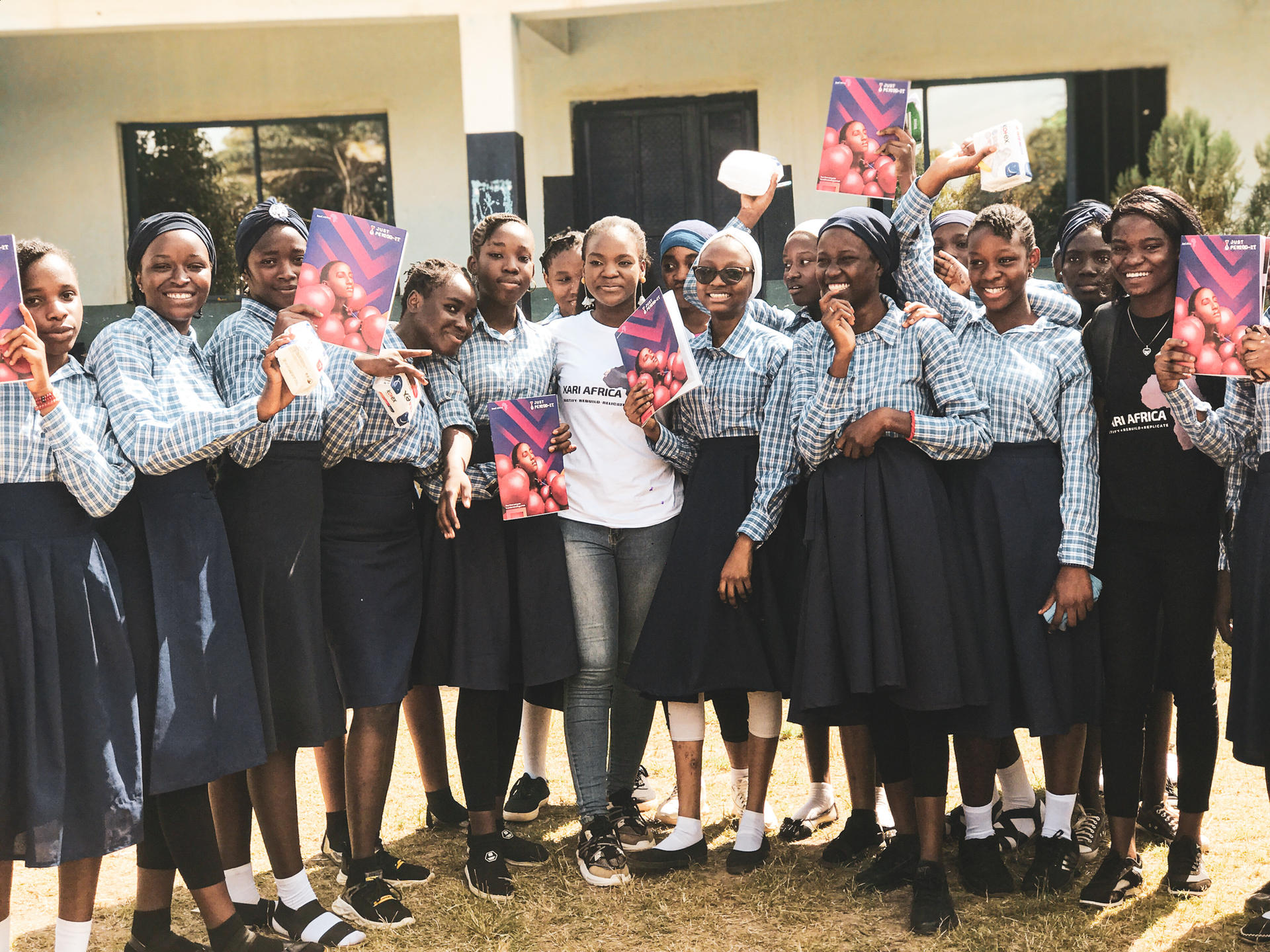 A group of smiling schoolgirls in uniforms poses with a woman, holding colorful materials outdoors.