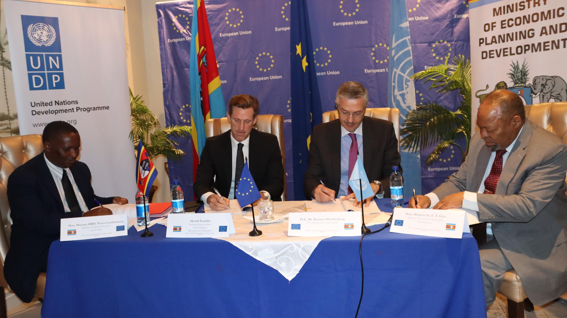 Four officials seated at a table signing documents, with flags and banners in the background.