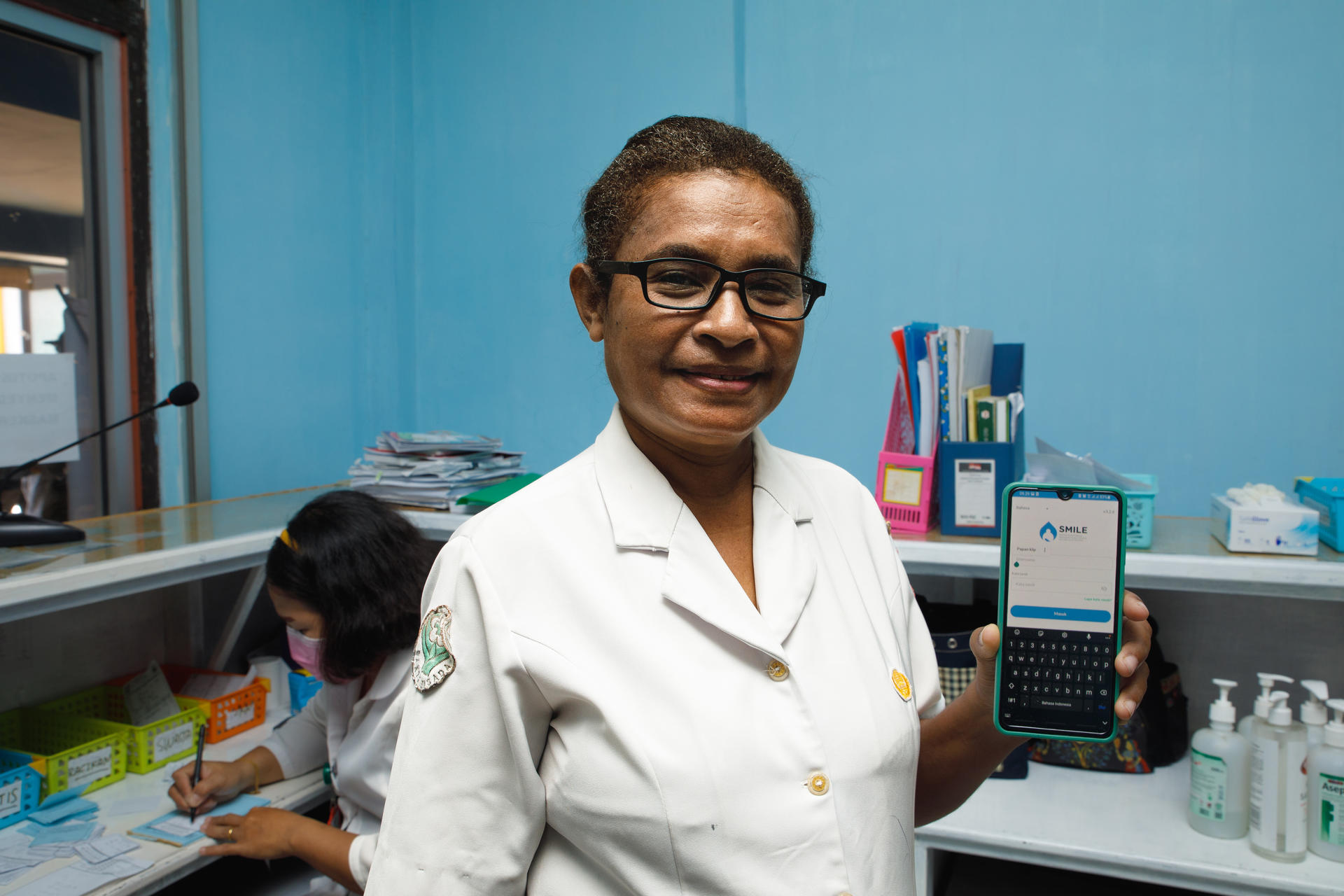 A nurse in a white uniform smiles while holding a smartphone, with a child writing in the background.