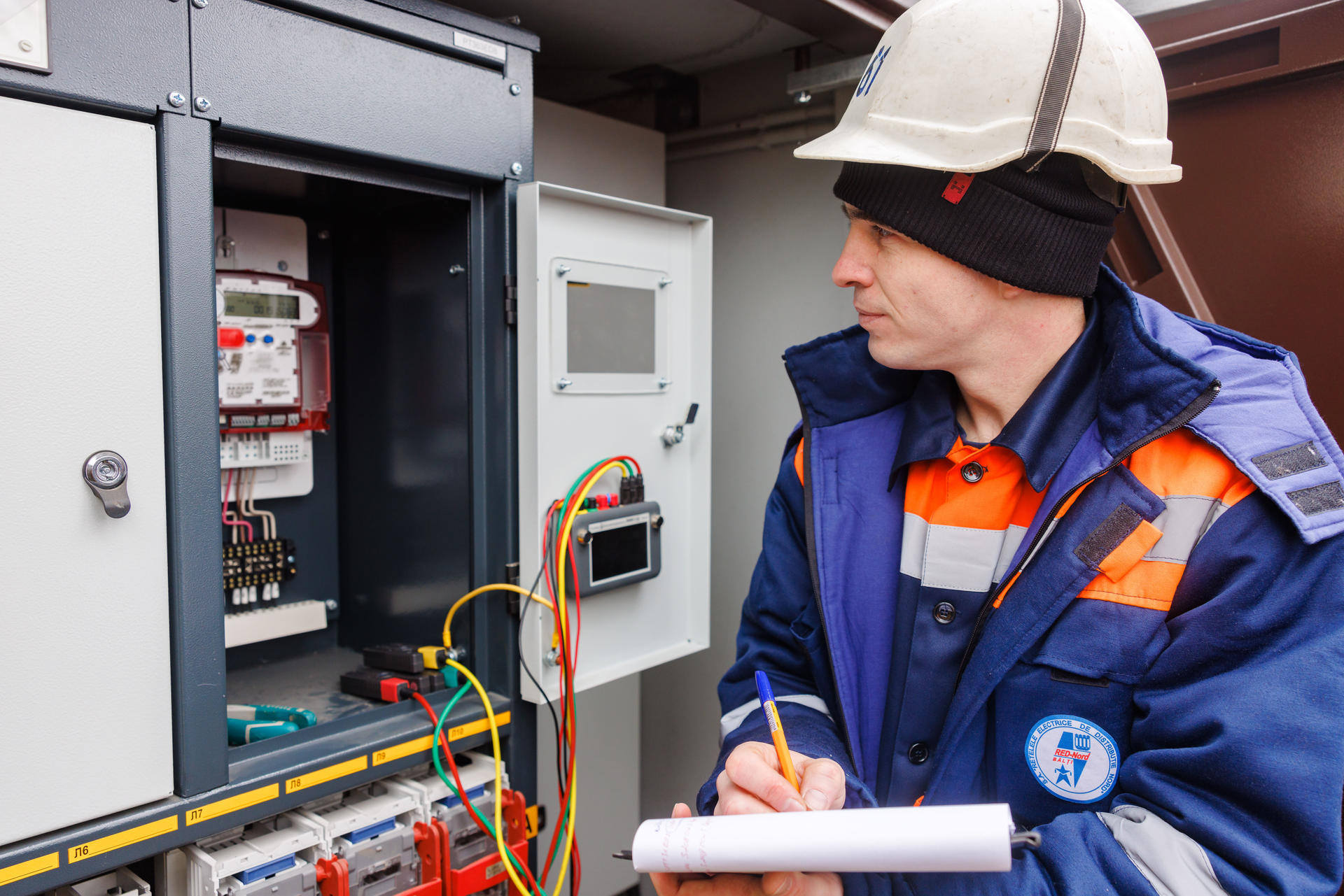 A technician in a hard hat examines an electrical panel with tools and notes.