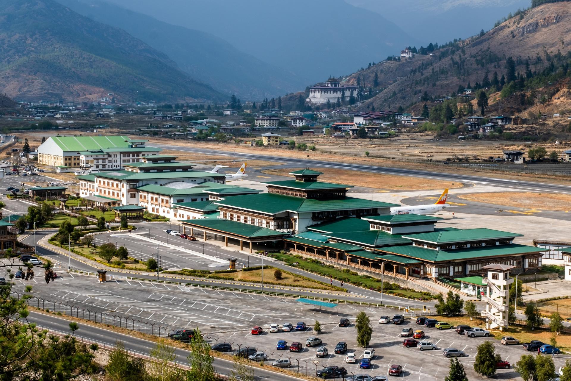 Aerial view of an airport terminal with green roofs, surrounded by mountains and a parking area.