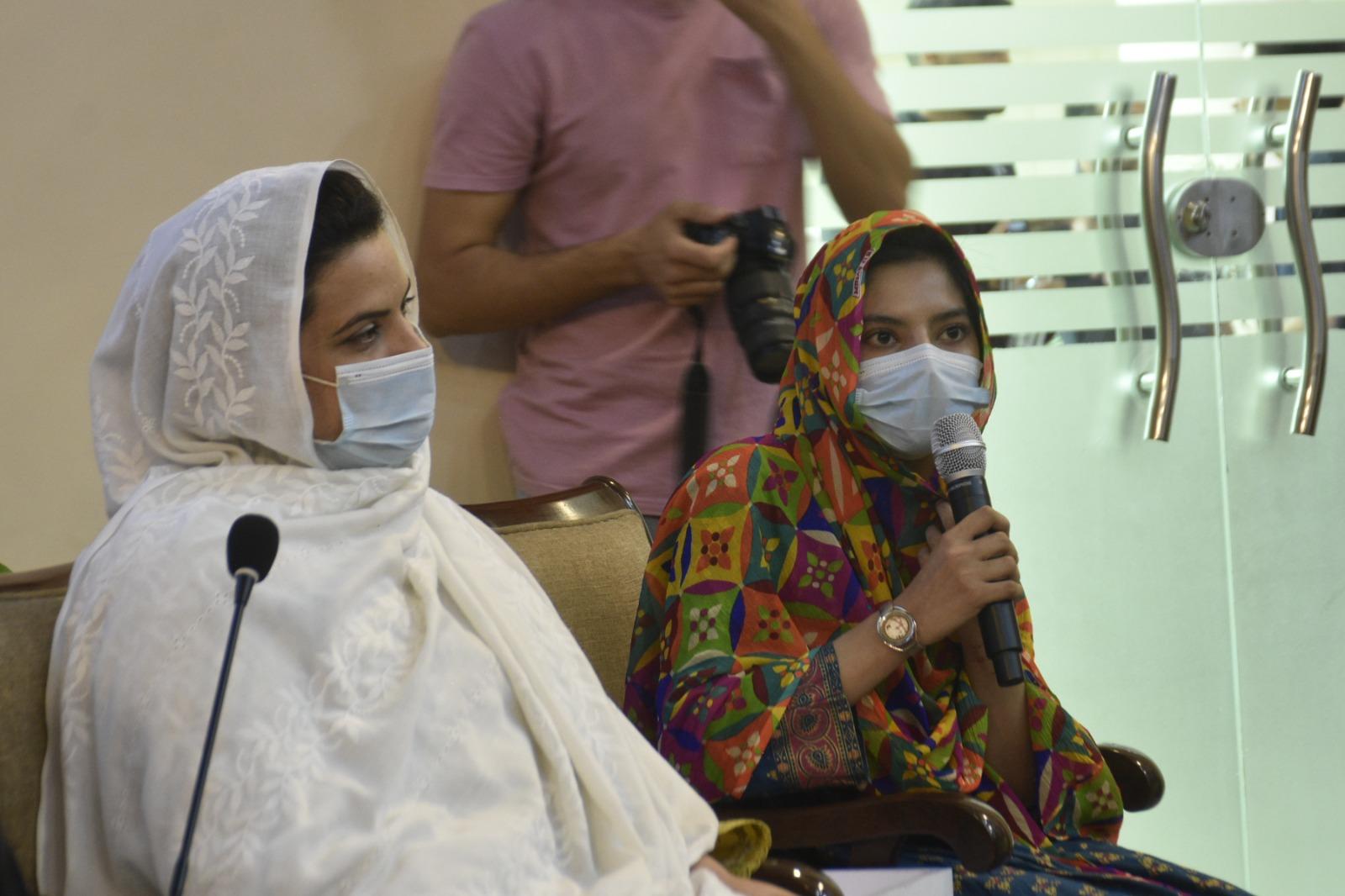 Two women sit in a conference room, one speaking into a microphone, both wearing masks.