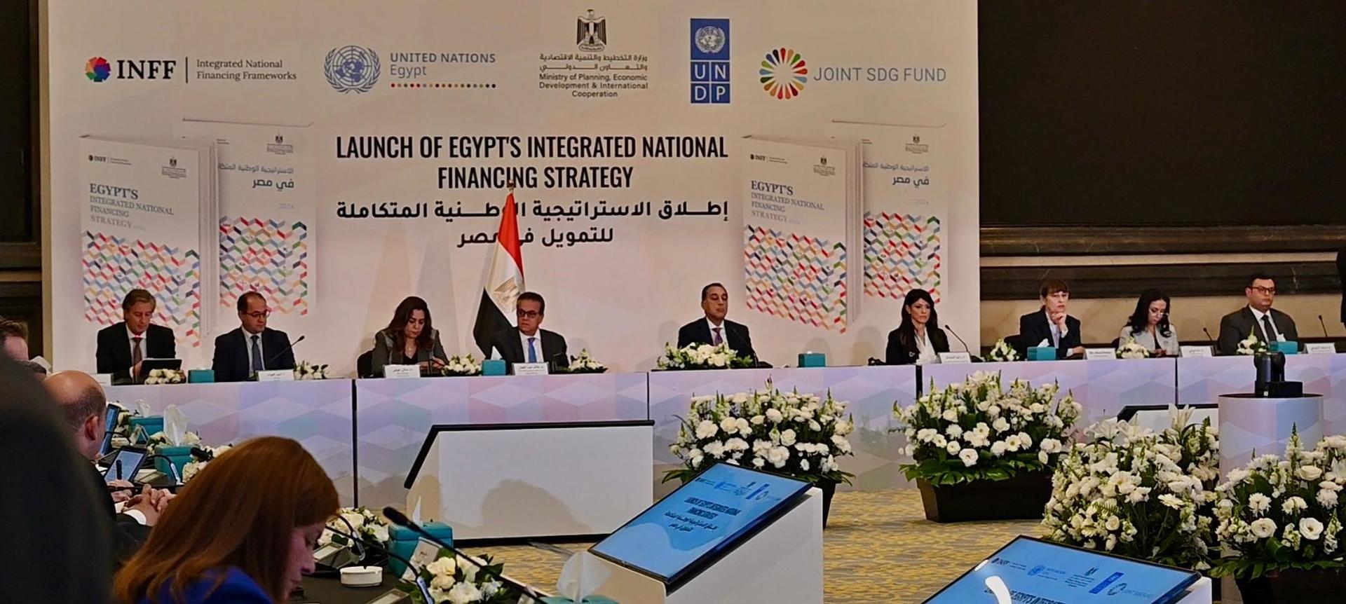 Conference setting with speakers at a table, national flags, and floral arrangements.